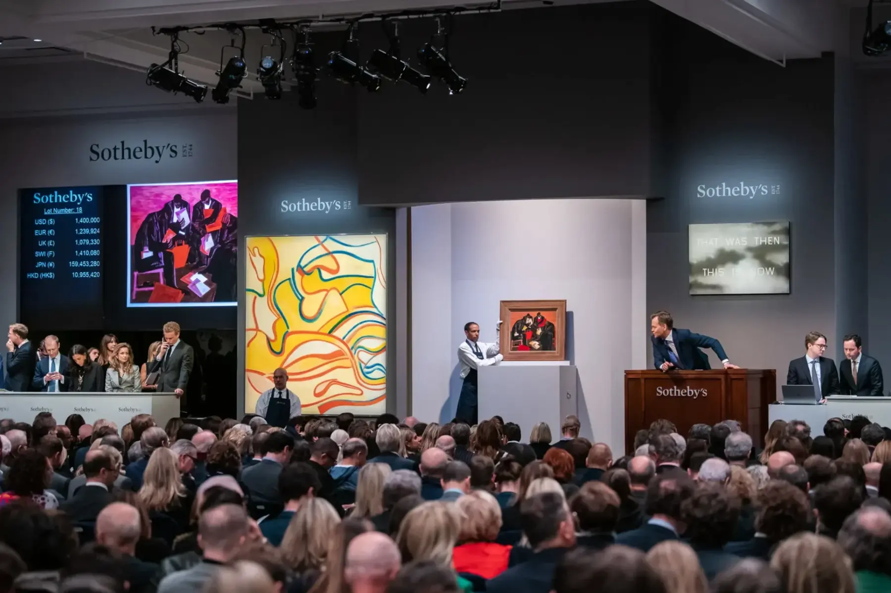 Crowded Sotheby’s auction with staff, auctioneer, and artwork displayed on walls and pedestal.