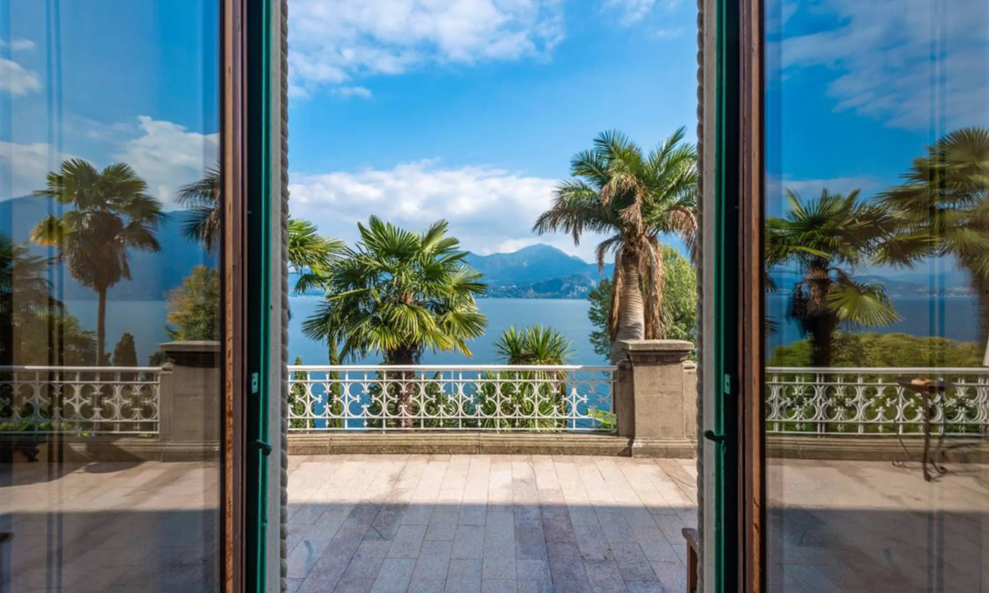View from open doors onto a terrace with palm trees and a lake with mountains under blue sky.