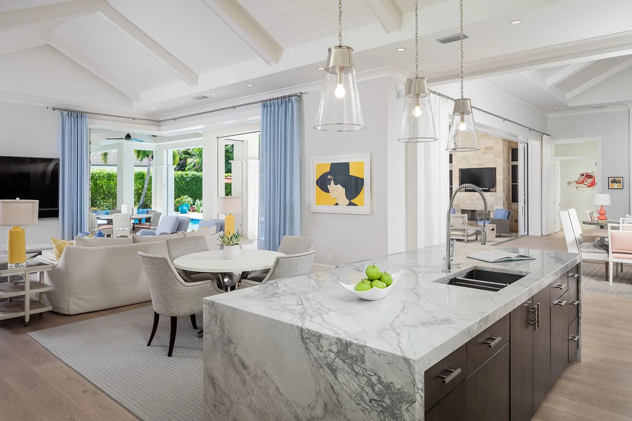 Blonde woman in blue suit smiling, standing in a modern living area with marble countertop and plants.