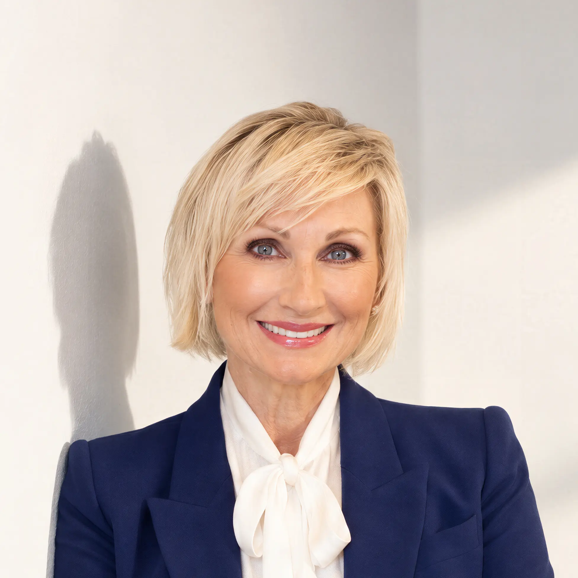 Smiling woman with short blonde hair wearing a blue blazer and white blouse bow tie.