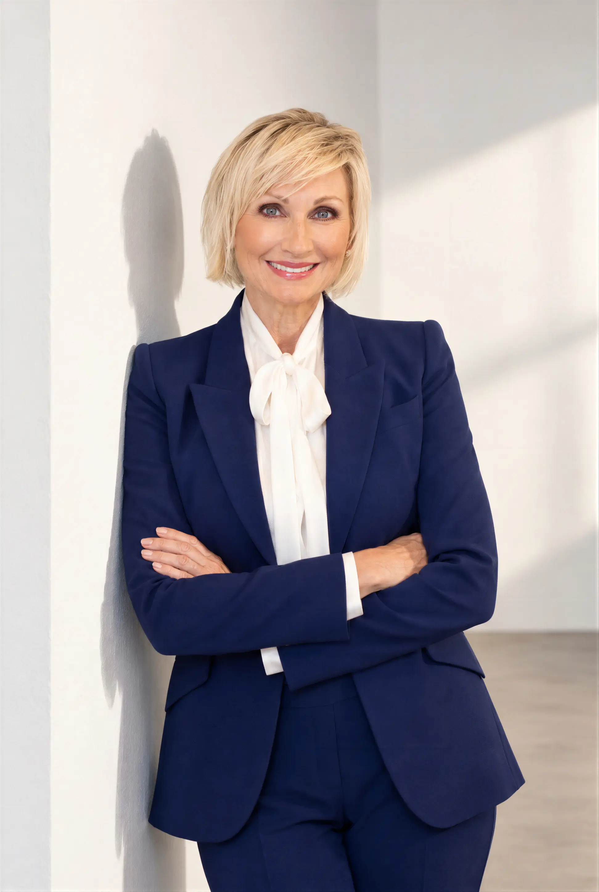 Blonde woman in blue suit smiling, standing in a modern living area with marble countertop and plants.