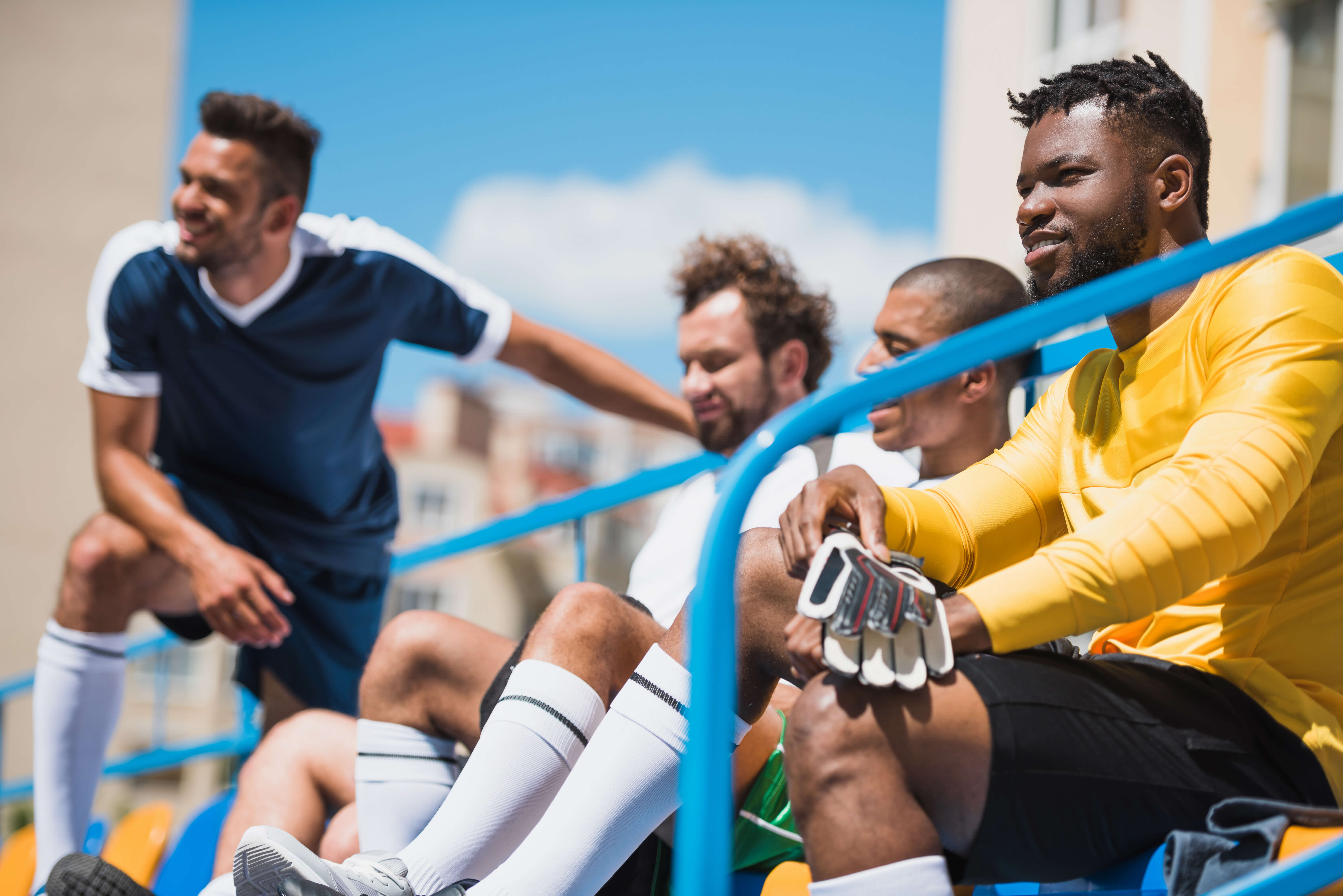 A multicultural group of male soccer players resting and sitting on stadium bleachers on a sunny day, one wearing a yellow goalkeeper jersey and gloves.  Lightning Performance Solutions