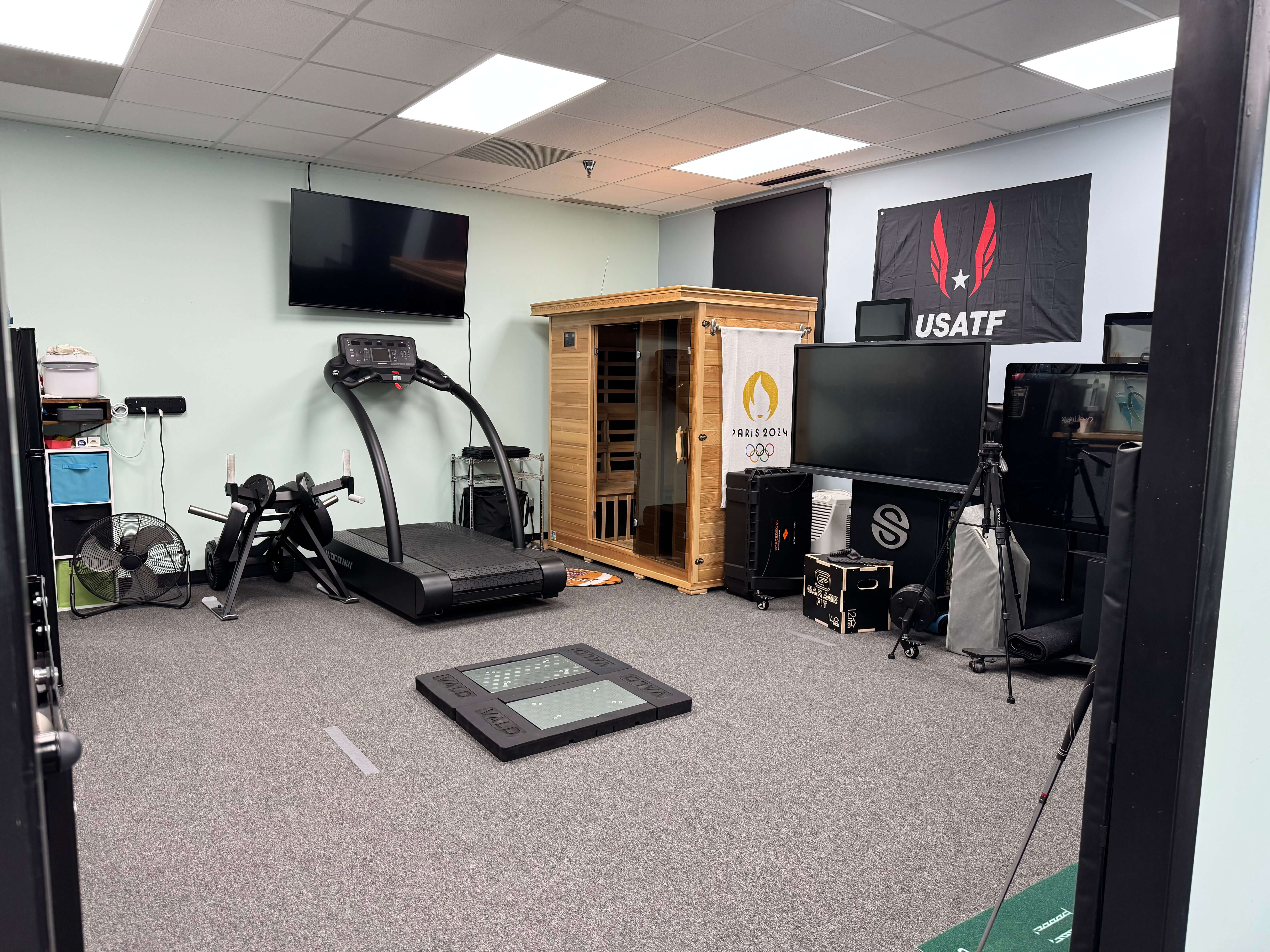 Indoor fitness room with treadmill, weight bench, wooden sauna, TV screens, fan, and USATF and Paris 2024 Olympic banners on the walls.  Lightning Performance Solutions