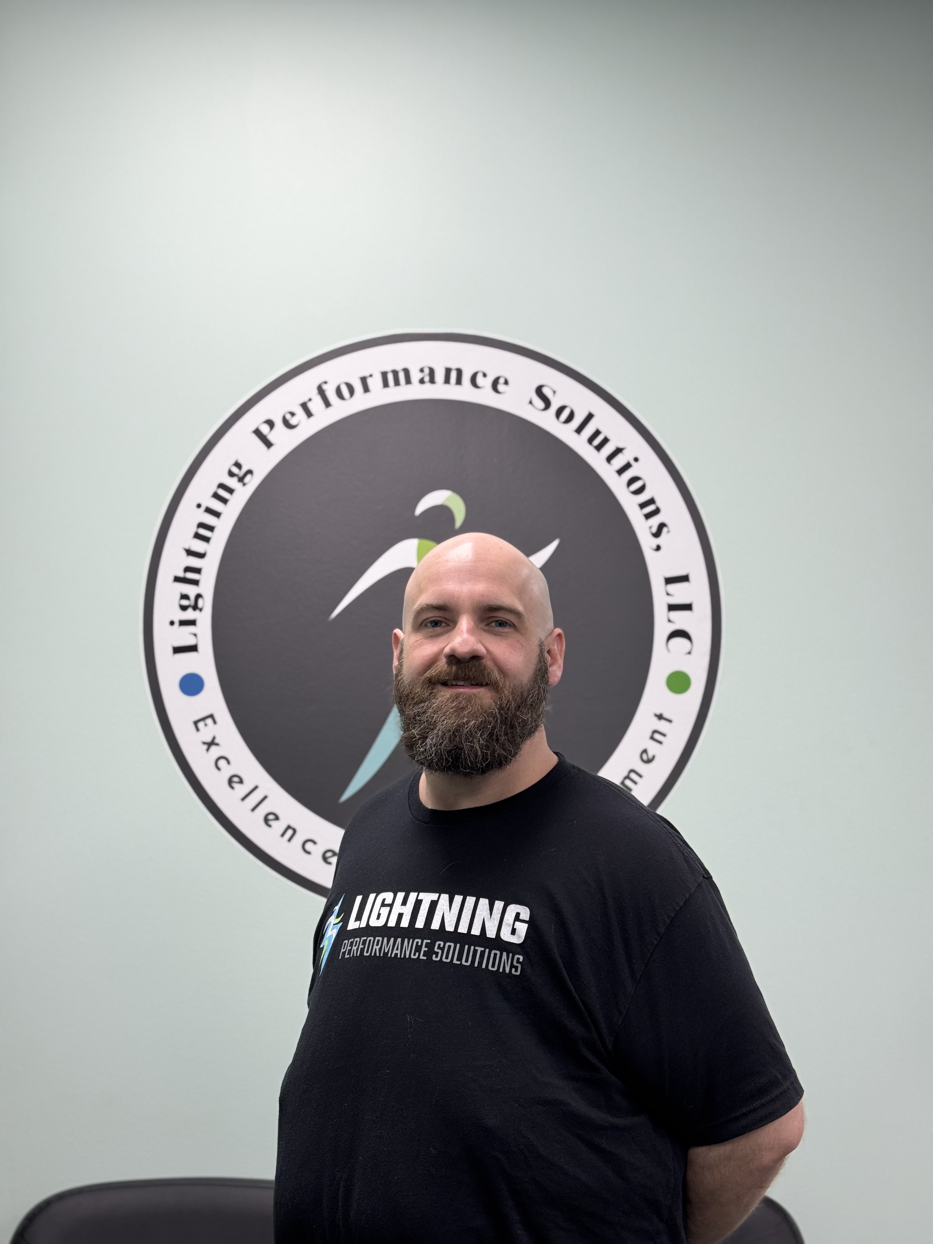 Bearded man wearing a black Lightning Performance Solutions t-shirt standing in front of a company logo on a light gray wall.  Lightning Performance Solutions