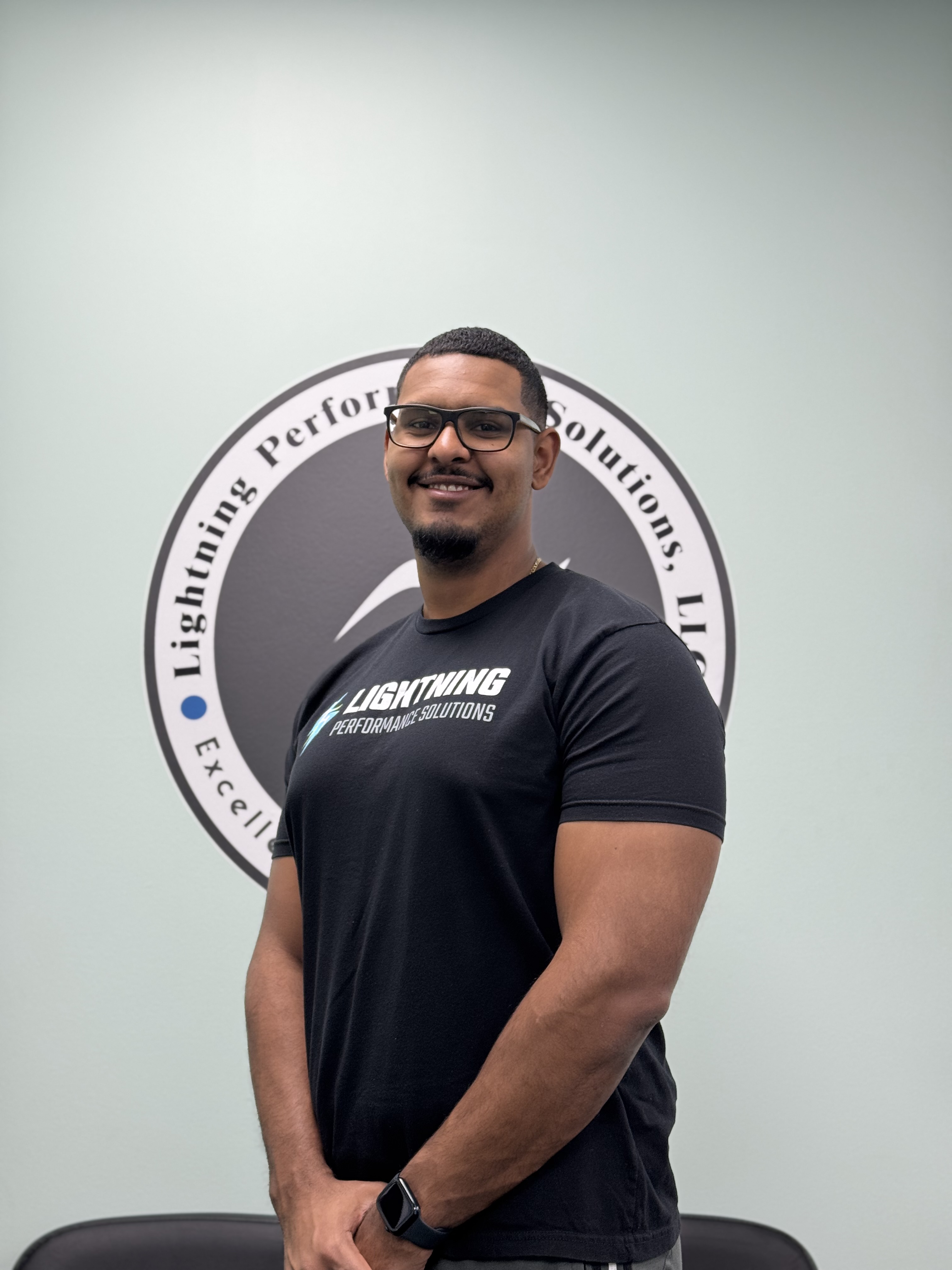 Smiling man wearing glasses and a black Lightning Performance Solutions t-shirt standing in front of a company logo on a light gray wall.  Lightning Performance Solutions