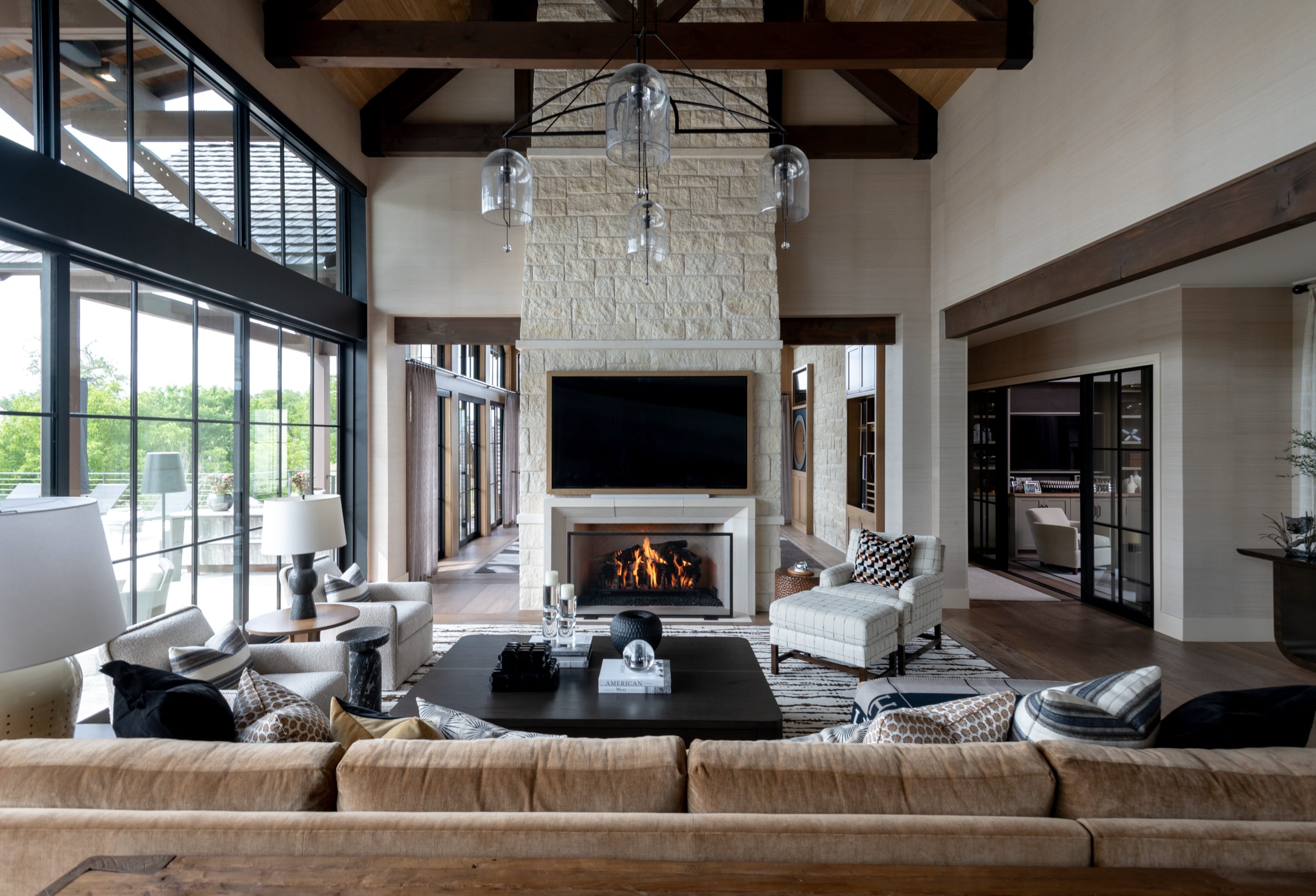 Spacious modern living room with large windows, beige sofa, armchairs, a fireplace with a flat-screen TV above, and wooden ceiling beams.
