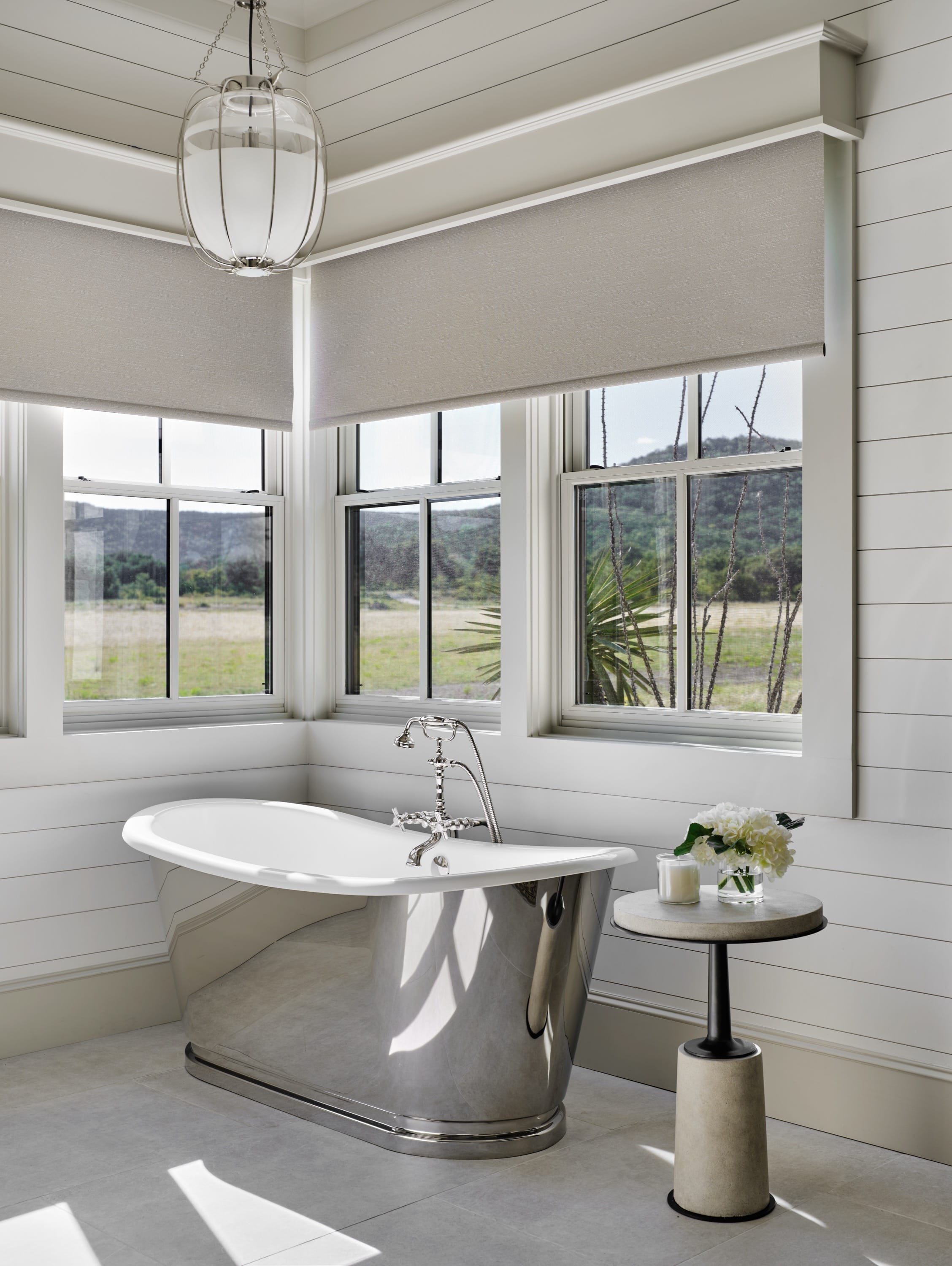 Modern bathroom corner with a reflective silver freestanding bathtub, three windows with beige blinds, and a small round table holding a candle and white flowers.