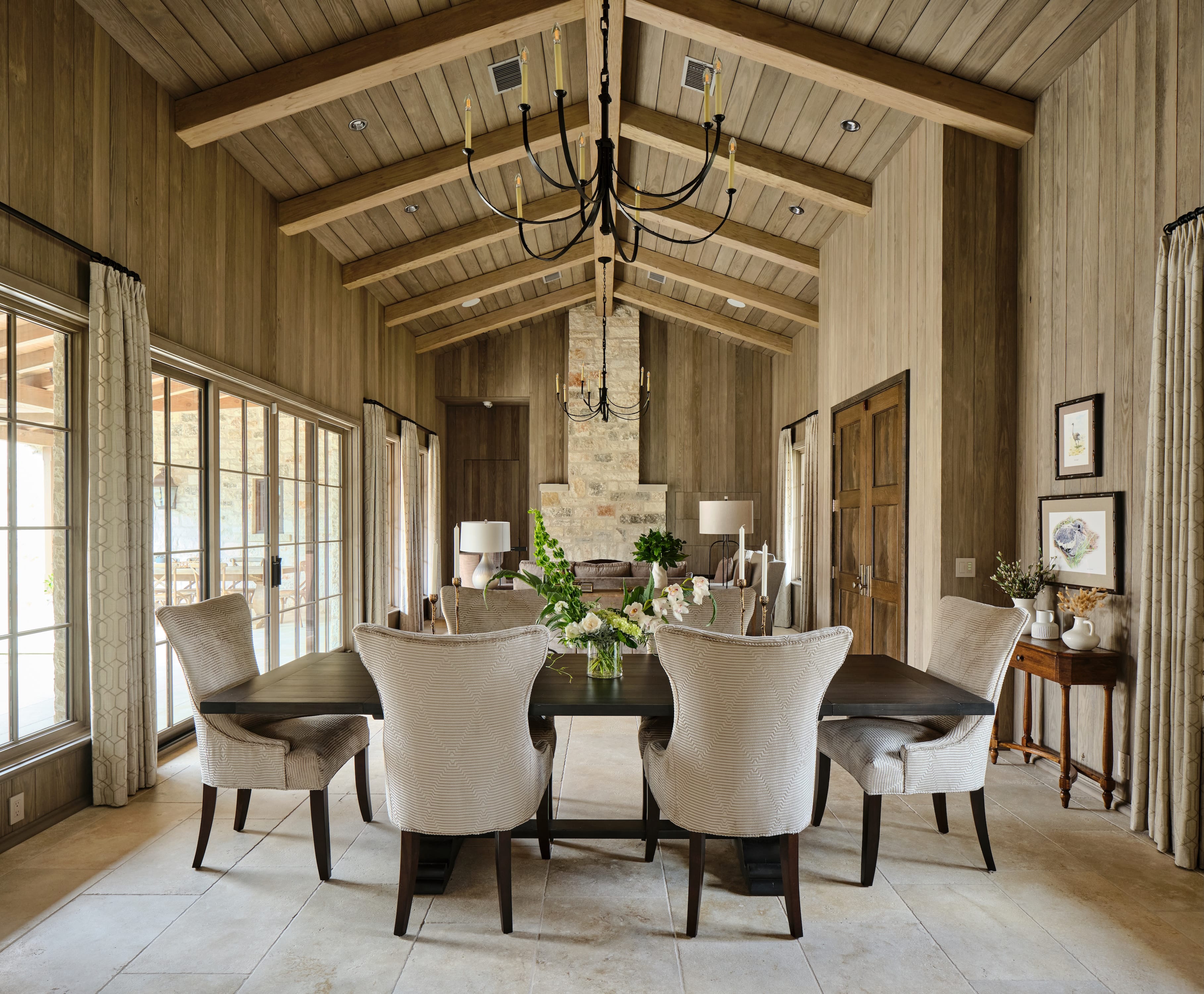 Dining room with a dark wooden table surrounded by six beige upholstered chairs, floral centerpiece, large windows with curtains, wooden walls and ceiling with exposed beams, and two chandeliers.