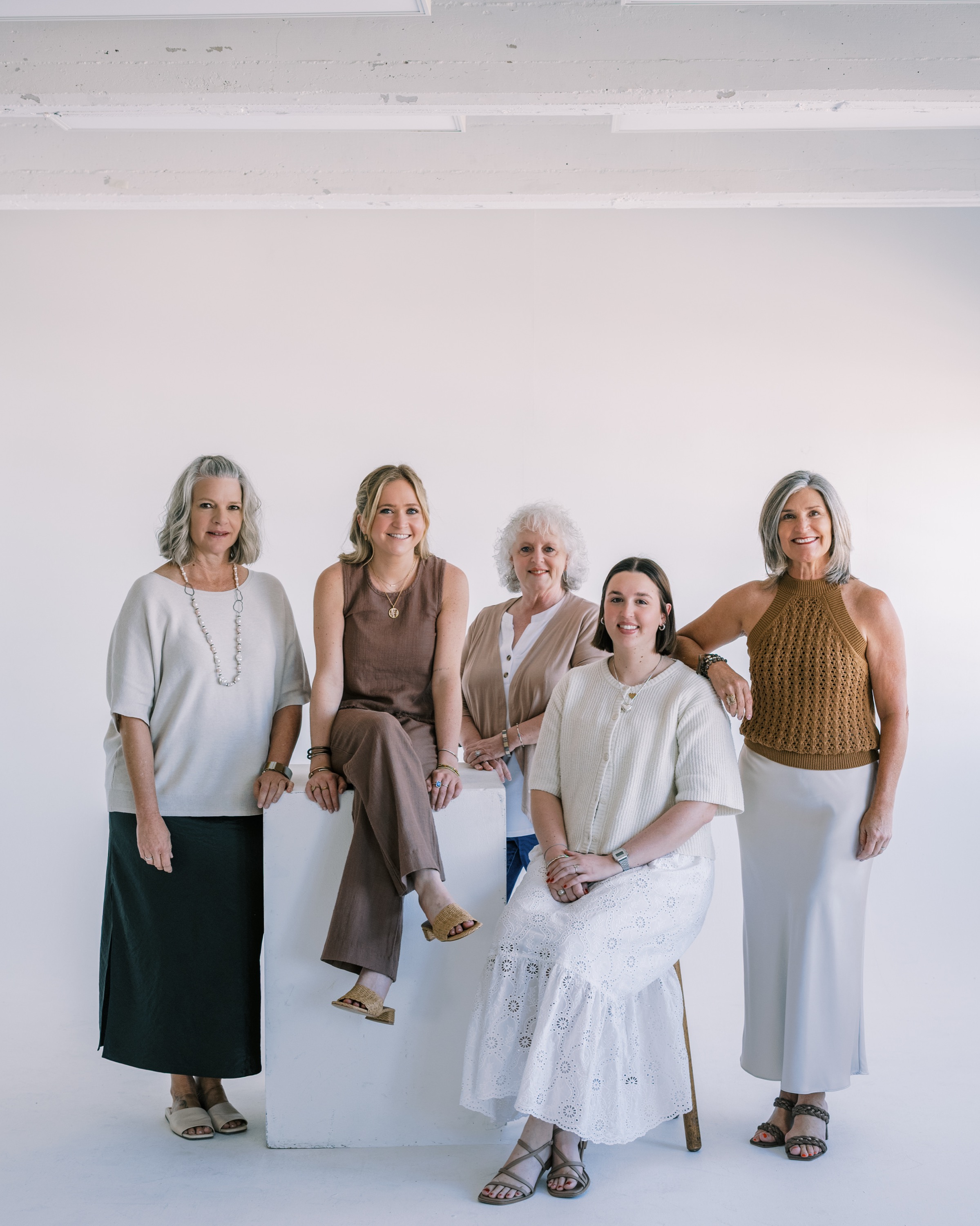 Five women posing against a white wall, dressed in neutral tones with casual and elegant outfits.
