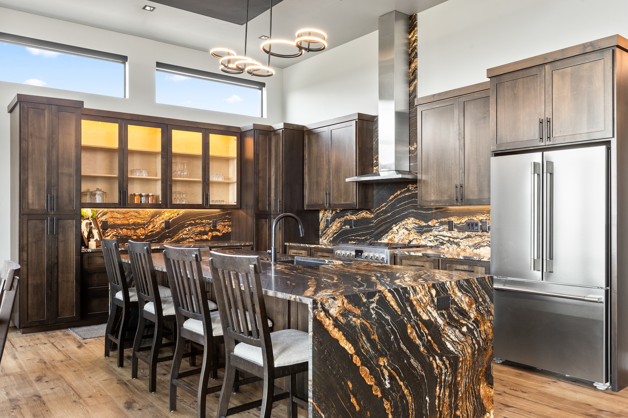 Modern kitchen with dark wood cabinets, a large island with black and orange marble countertop and backsplash, stainless steel refrigerator, and pendant lighting.