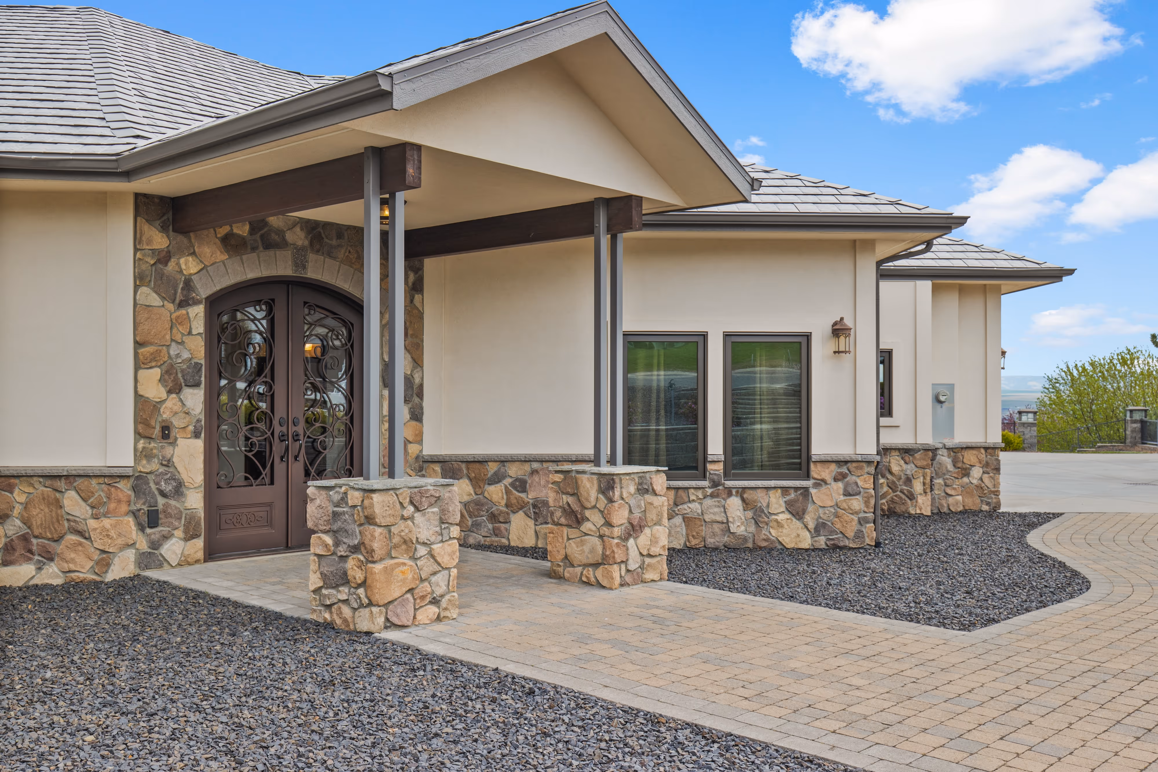 Modern house entrance with double decorative wrought iron doors, stone and stucco exterior, and paved walkway.