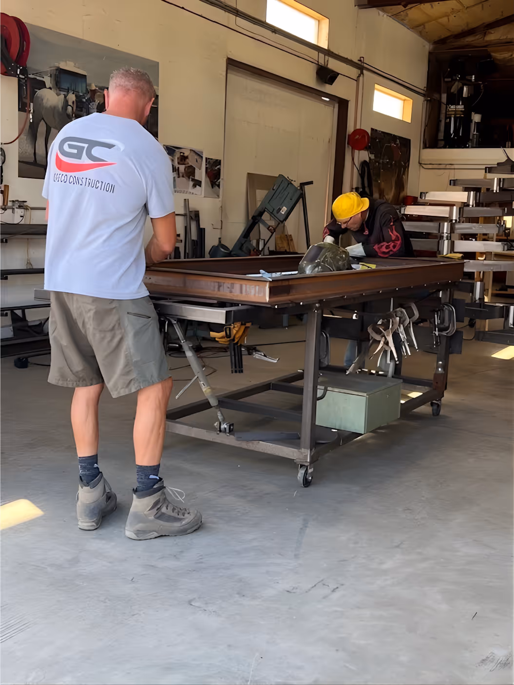 Two men working together at a large metal table in a workshop, one wearing a yellow hard hat and the other in a light blue shirt and gray shorts.