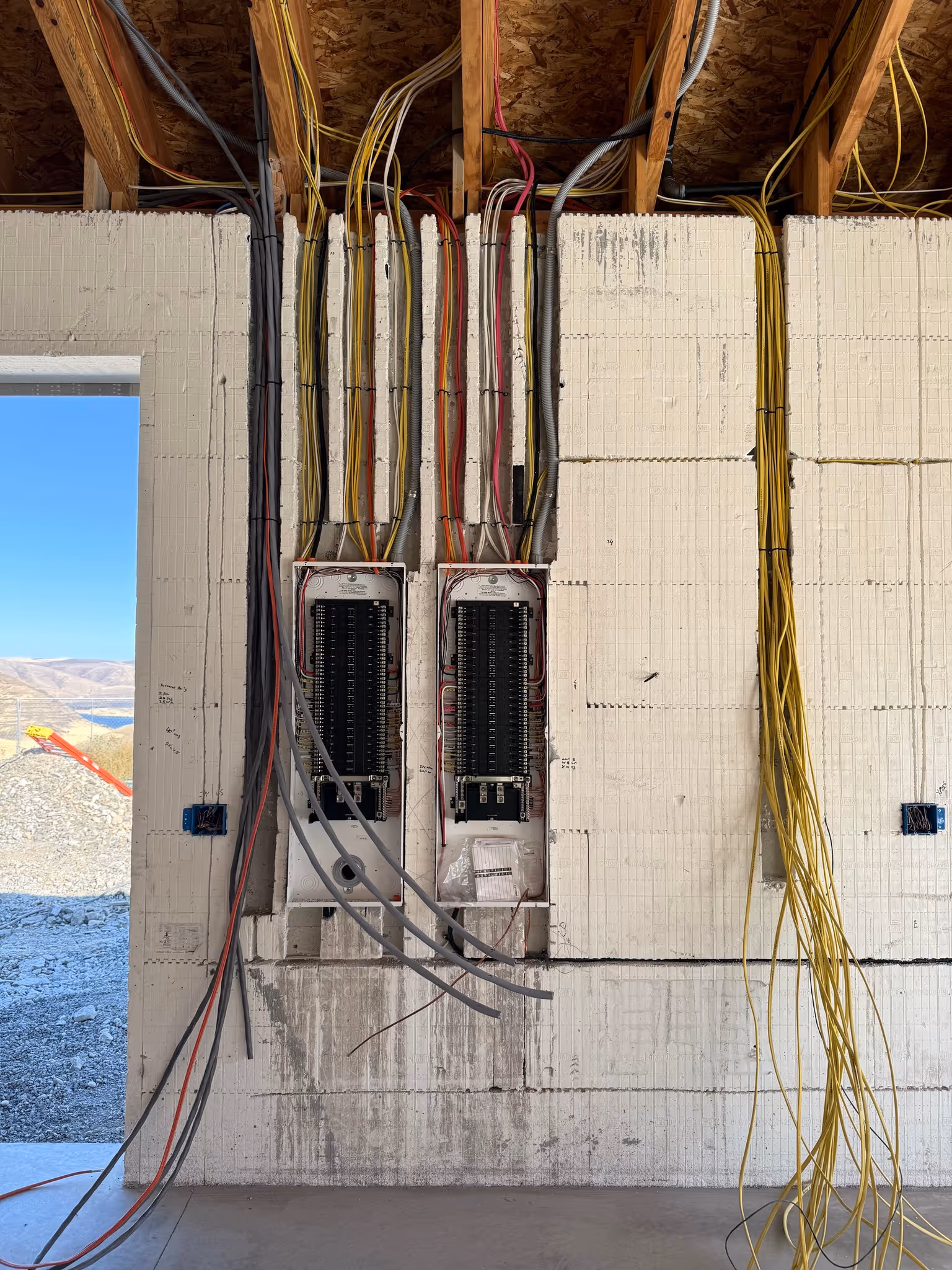 Two electrical panels installed in a wall under construction with various colored wires running vertically and bundled, and an open doorway to a rocky outdoor landscape.
