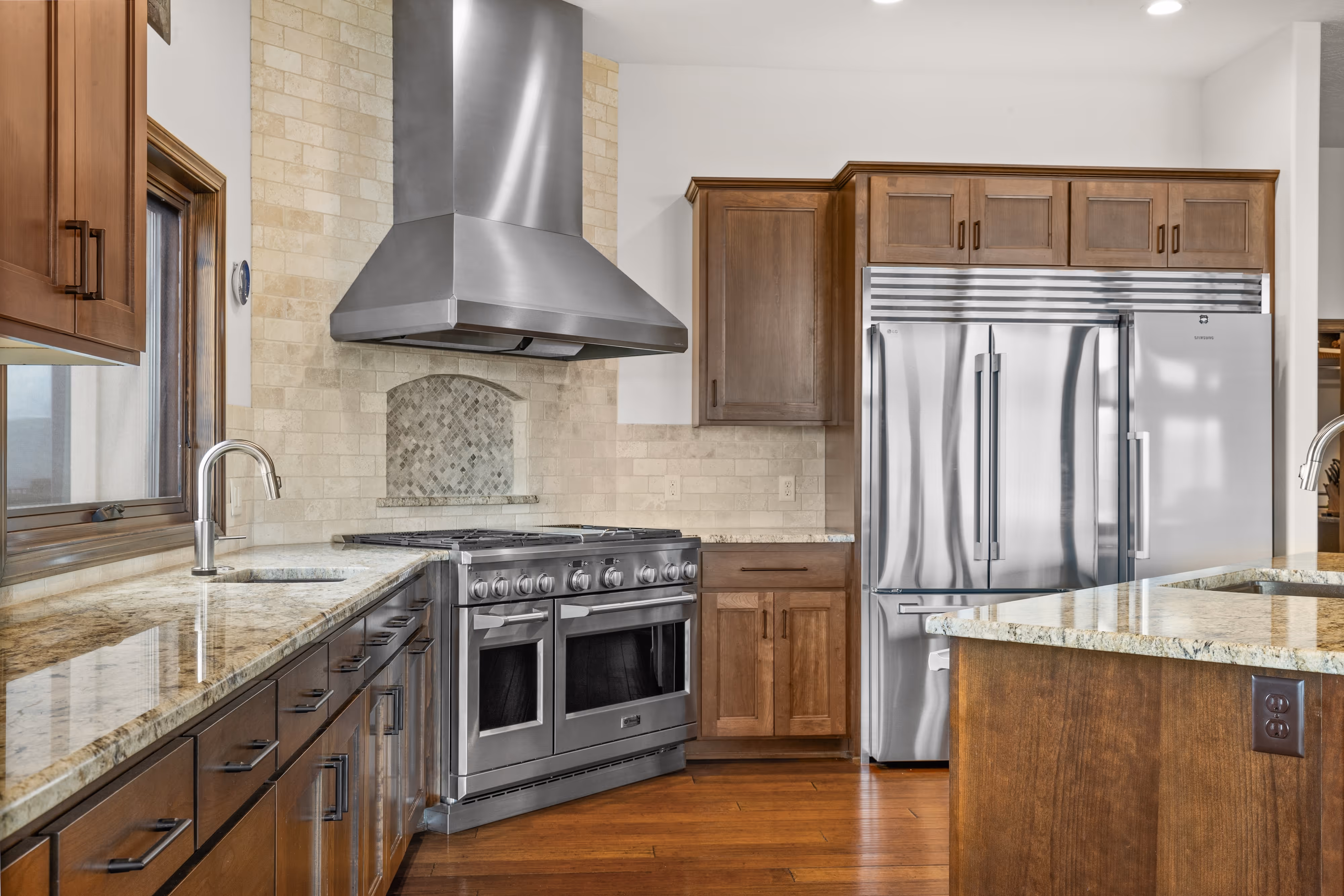 Modern kitchen with stainless steel range hood, large stove, double-door refrigerator, wood cabinets, and granite countertops.