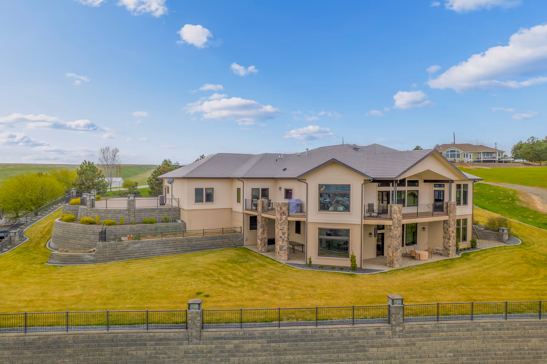 Modern two-story house with beige exterior, stone pillars, large windows, and tiered retaining walls on a grassy lawn under a blue sky.