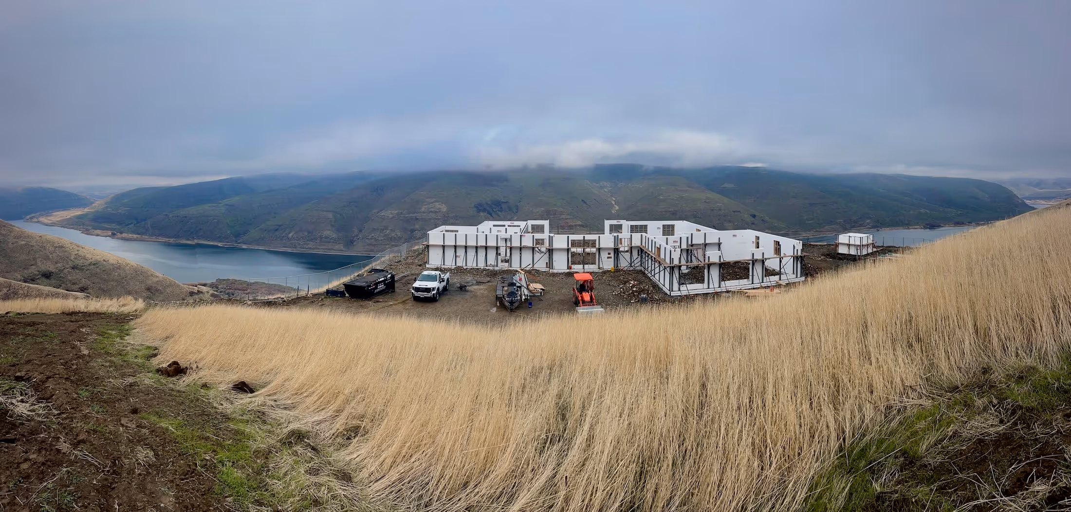 Construction site of a large building with white walls on a hillside overlooking a river and mountains under a cloudy sky.