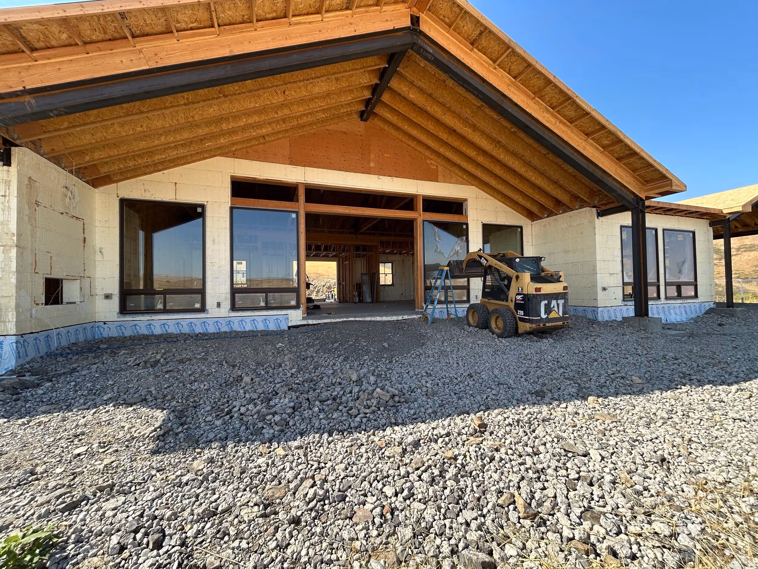 Partially constructed house with exposed wooden roof beams, large windows, a gravel yard, and a small CAT construction loader and ladder in front.