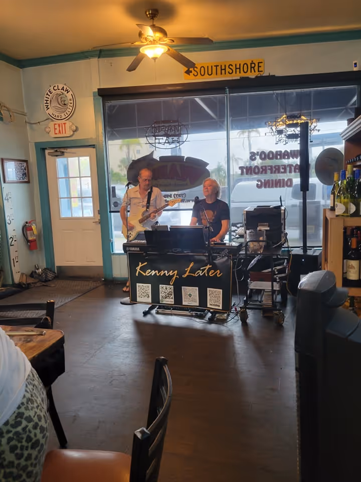 Two musicians perform in a cozy indoor setting, one plays guitar and the other keyboard. A "Kenny Later" sign is displayed. The mood is relaxed.