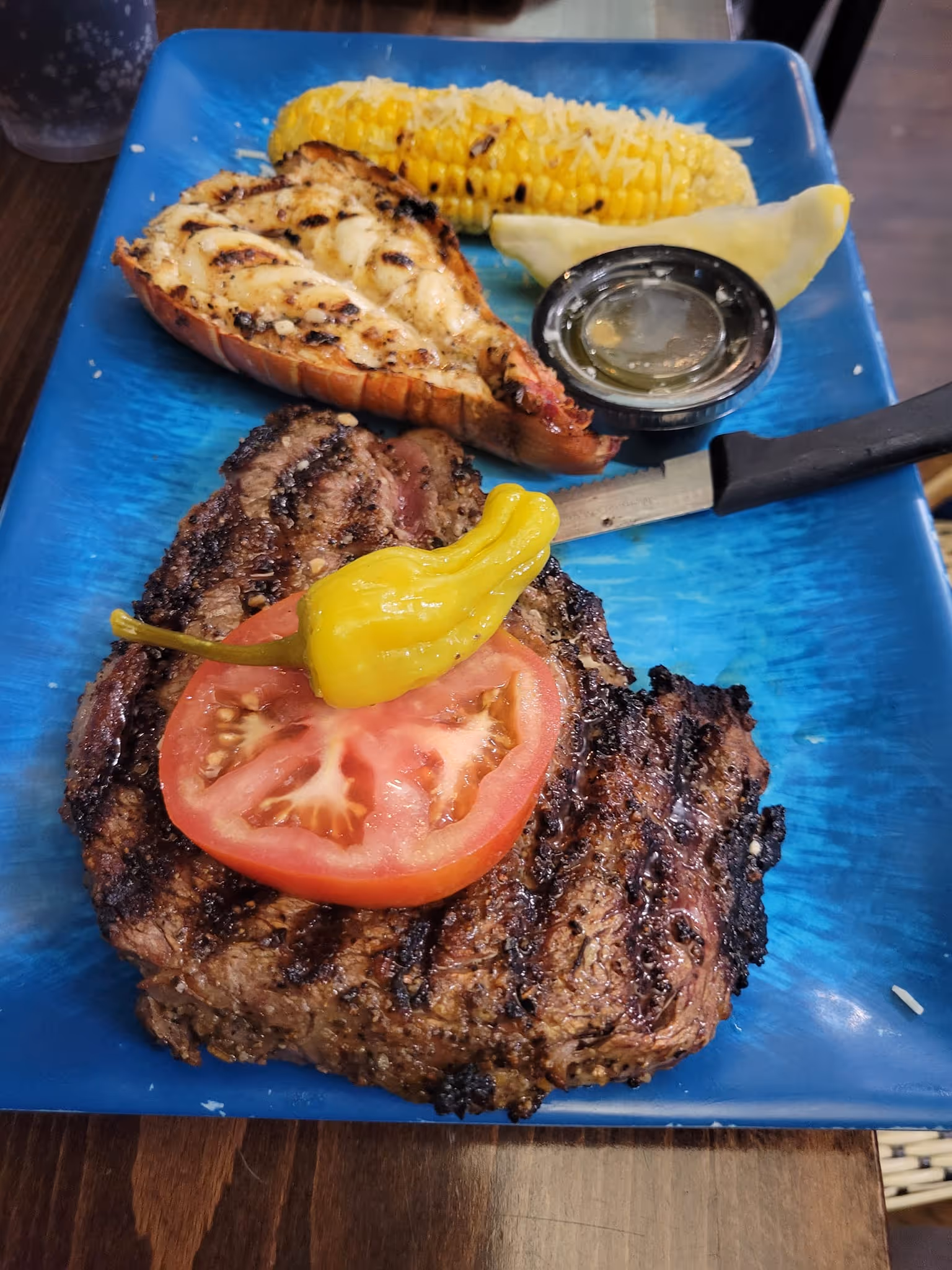 Grilled steak topped with tomato and pepper, served with a lobster tail, corn on the cob, and lemon wedge on a blue plate; savory and appetizing.