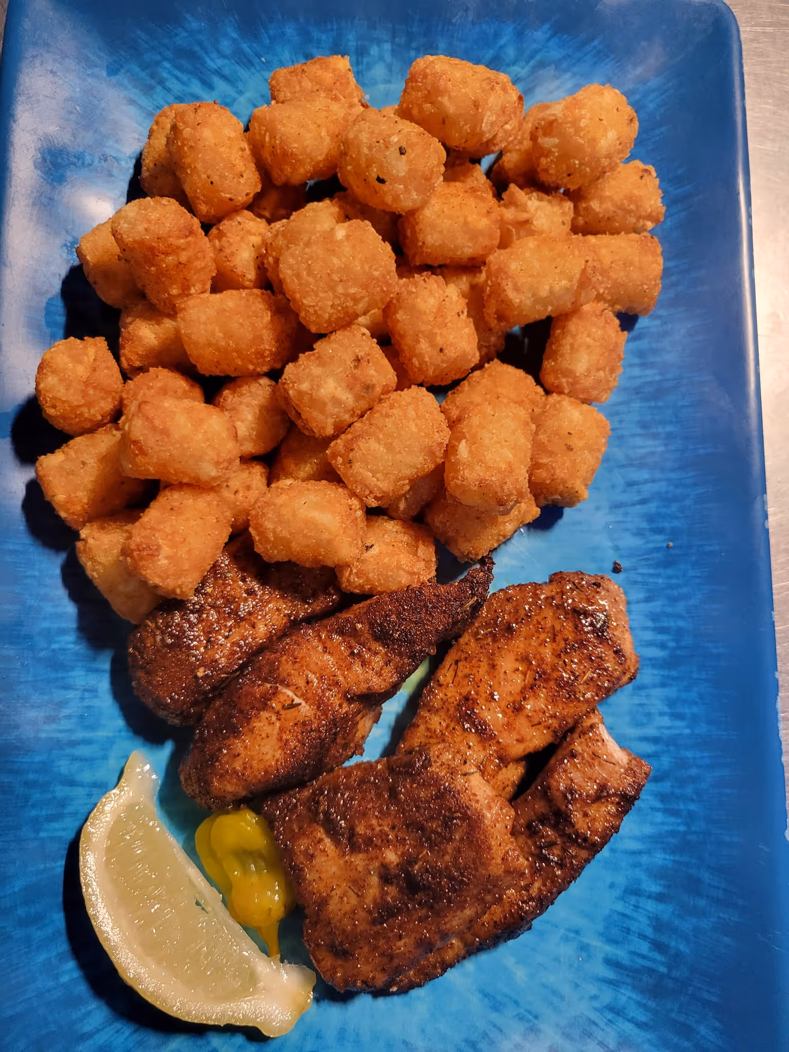 A blue plate features a mound of golden-brown tater tots, blackened meat slices, a lemon wedge, and a yellow pepper, evoking a cozy, inviting meal.