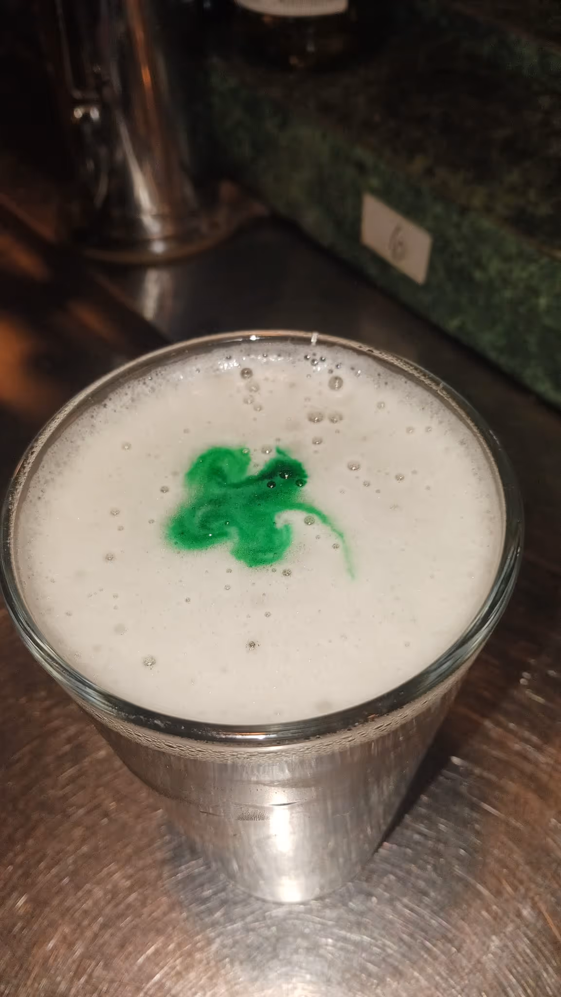 A pint of beer with creamy white foam topped with a green shamrock design. The setting suggests a casual and festive St. Patrick's Day mood.