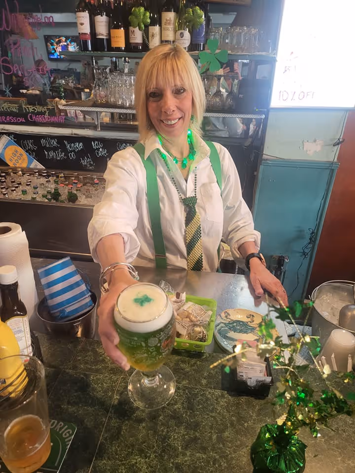 A smiling bartender wearing green suspenders and a tie offers a green beer with a shamrock design. The festive bar is decorated for St. Patrick’s Day.