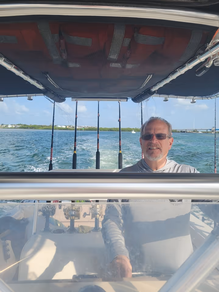 A man in sunglasses steering a boat on open water under a sunny sky. Fishing rods are mounted on the boat, and the mood is relaxed and adventurous.