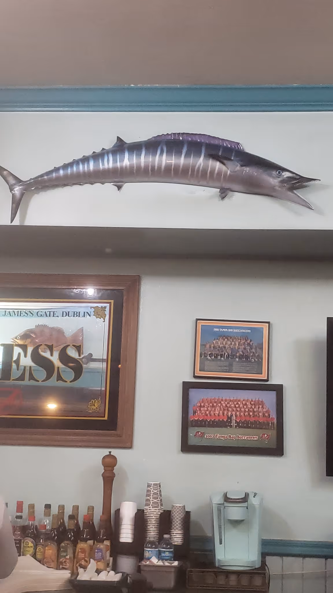 Wall with a mounted fish, framed photos, and a Dublin sign. Beneath are bottles, cups, and a coffee maker, creating a cozy, pub-like atmosphere.