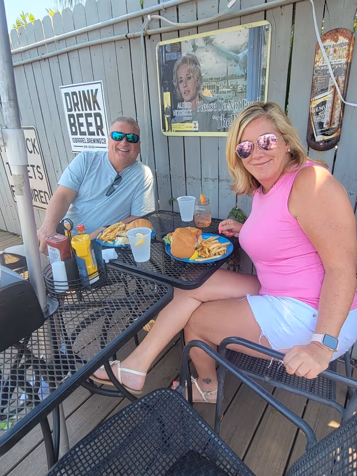 A man and woman enjoying a casual outdoor meal on a sunny day. They sit at a black metal table with burgers, fries, and drinks. Both are smiling, and the atmosphere is relaxed and cheerful. A wooden fence with vintage-style posters and a "Drink Beer" sign is in the background.