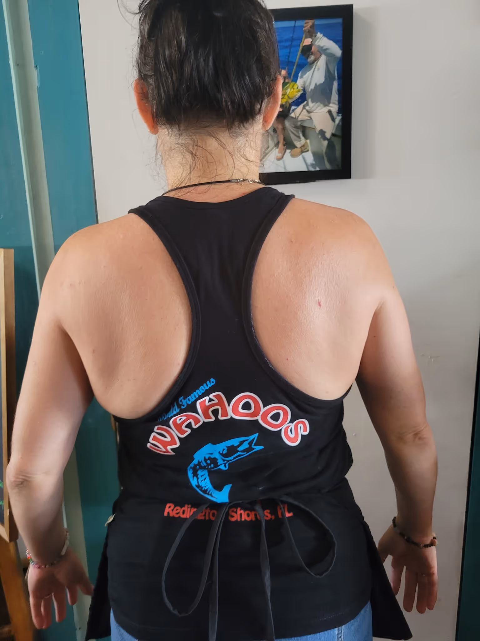 Rear view of a person with dark hair in a bun wearing a black "Wahoos" tank top featuring a blue fish logo. A framed photo is partially visible on the wall.