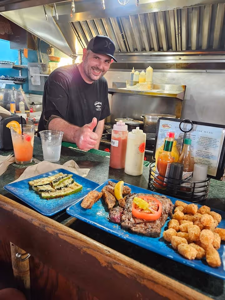 A cheerful cook gives a thumbs-up behind a counter with blue plates of grilled steak, zucchini, and tater tots. Condiments and cocktails are nearby.