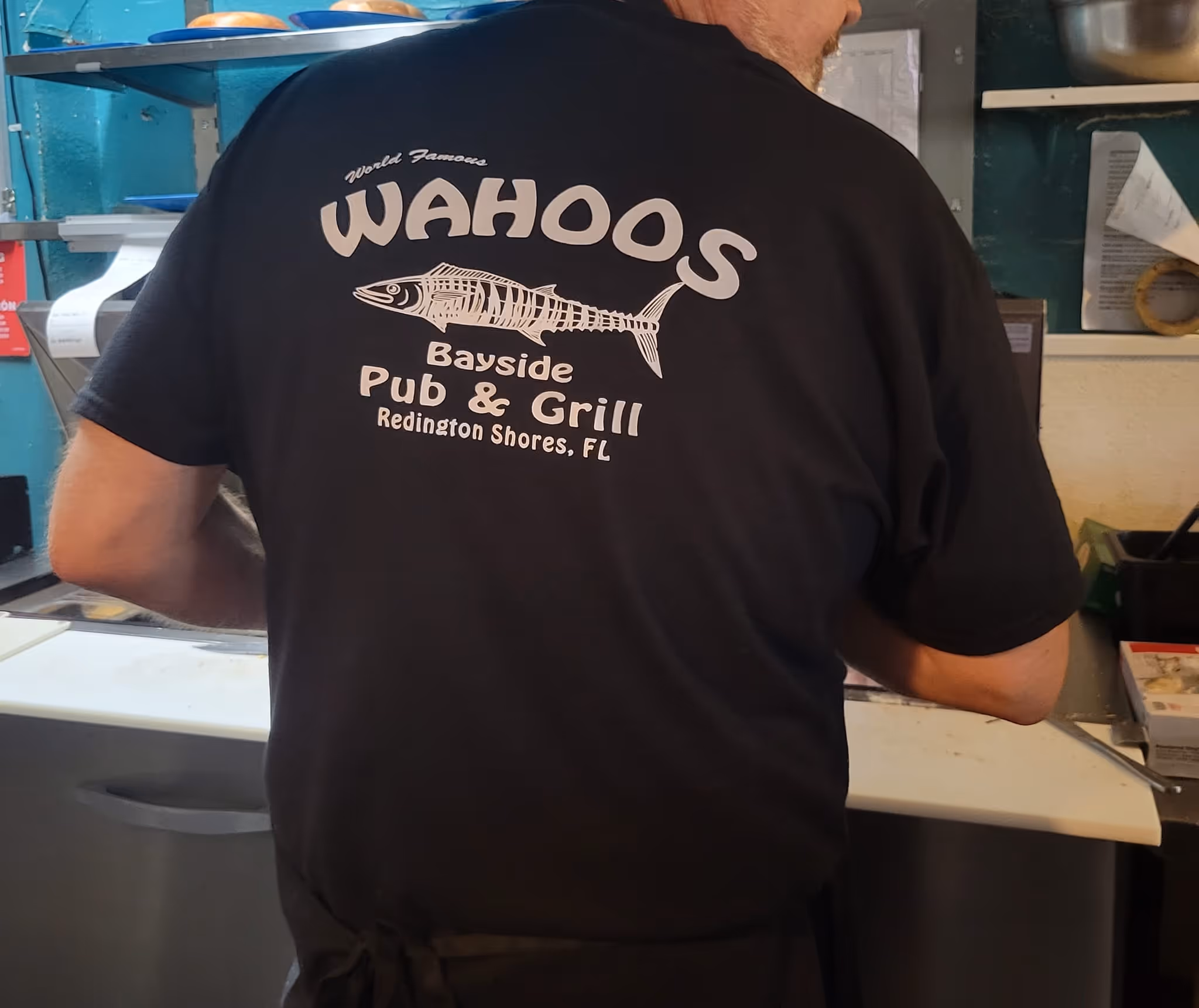 A person wears a black "Wahoos Pub & Grill" shirt with a fish design, standing in a kitchen setting, evoking a casual, local dining atmosphere.