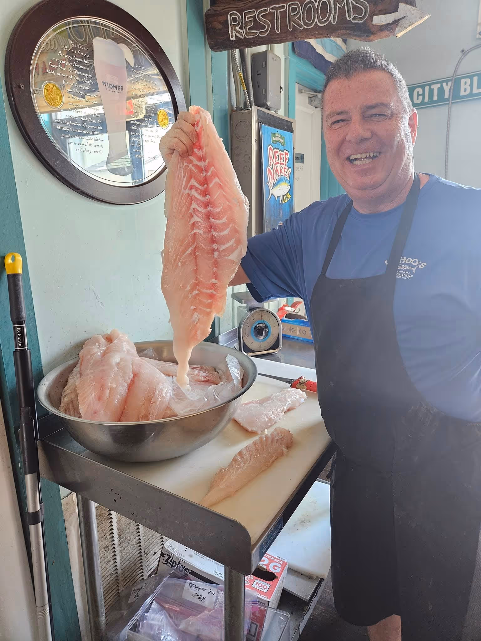 A person in an apron and blue shirt happily holds up a large, raw fish fillet over a metal bowl inside a shop. The room has a rustic style, featuring a "restrooms" sign and beer advertisements.