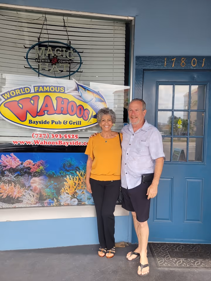 A smiling couple stands in front of Wahoo's Bayside Pub & Grill. The woman wears a yellow top; the man in a light shirt. Background: colorful sign and blue door.