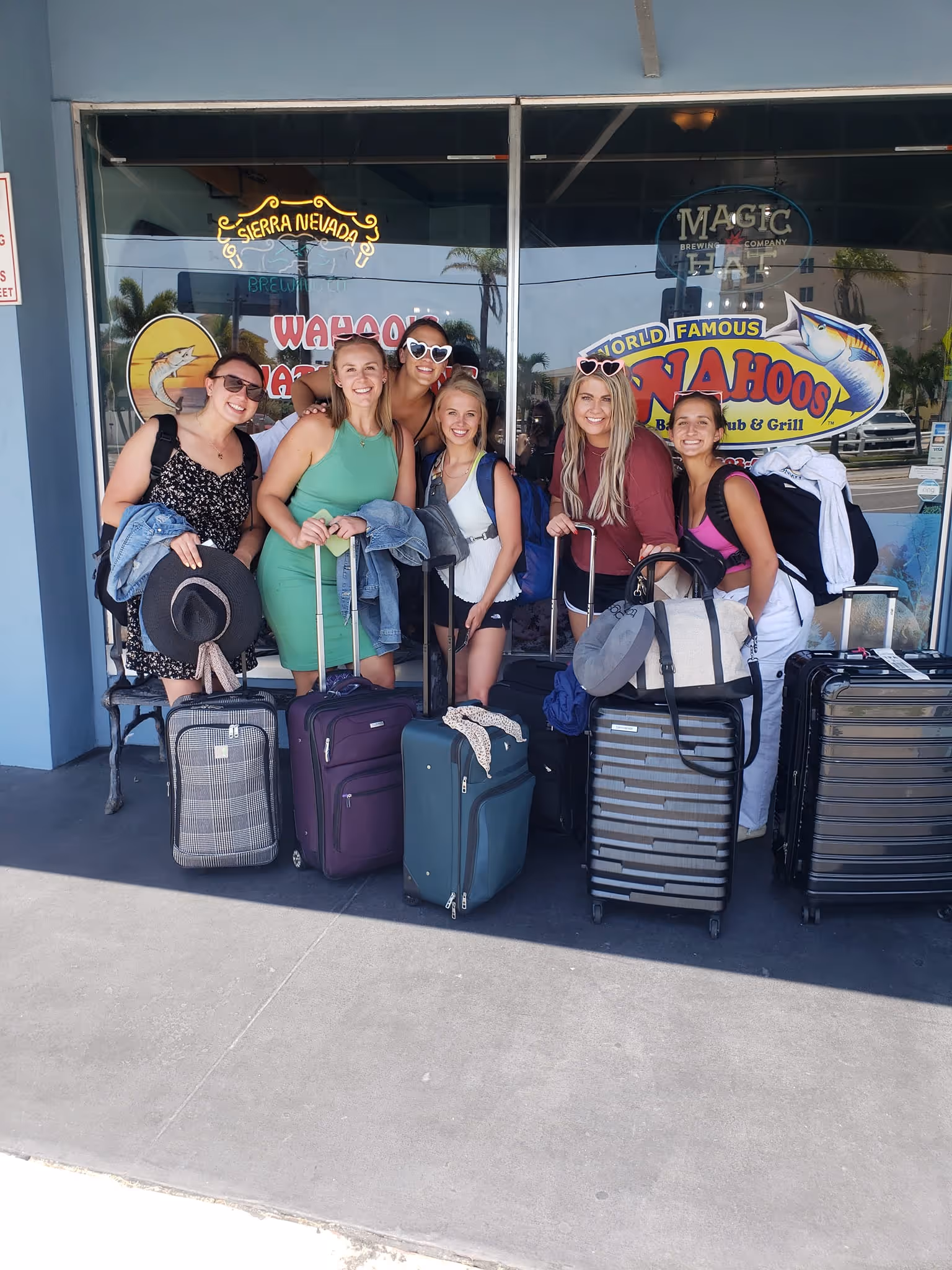 A group of six friends smiles outside a restaurant with luggage, suggesting travel. They appear joyful and ready for an adventure, featuring colorful outfits.