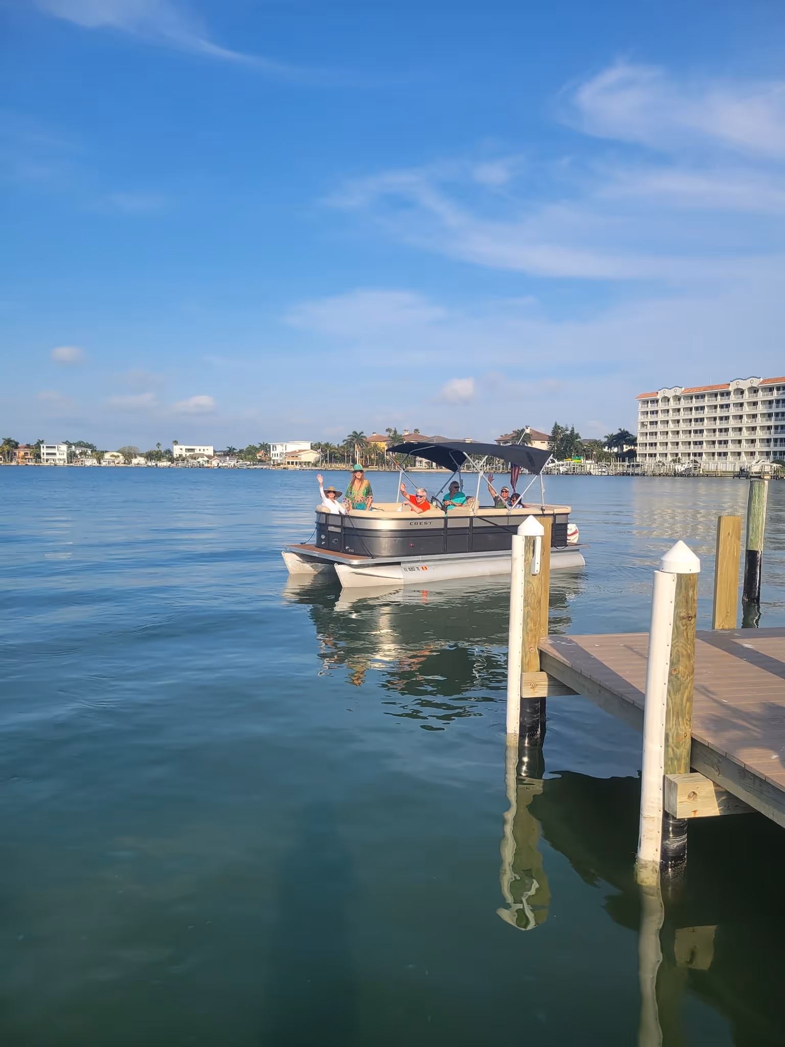 A pontoon boat filled with people enjoys a sunny day on calm waters near a wooden dock. Nearby are waterfront buildings under a clear blue sky.