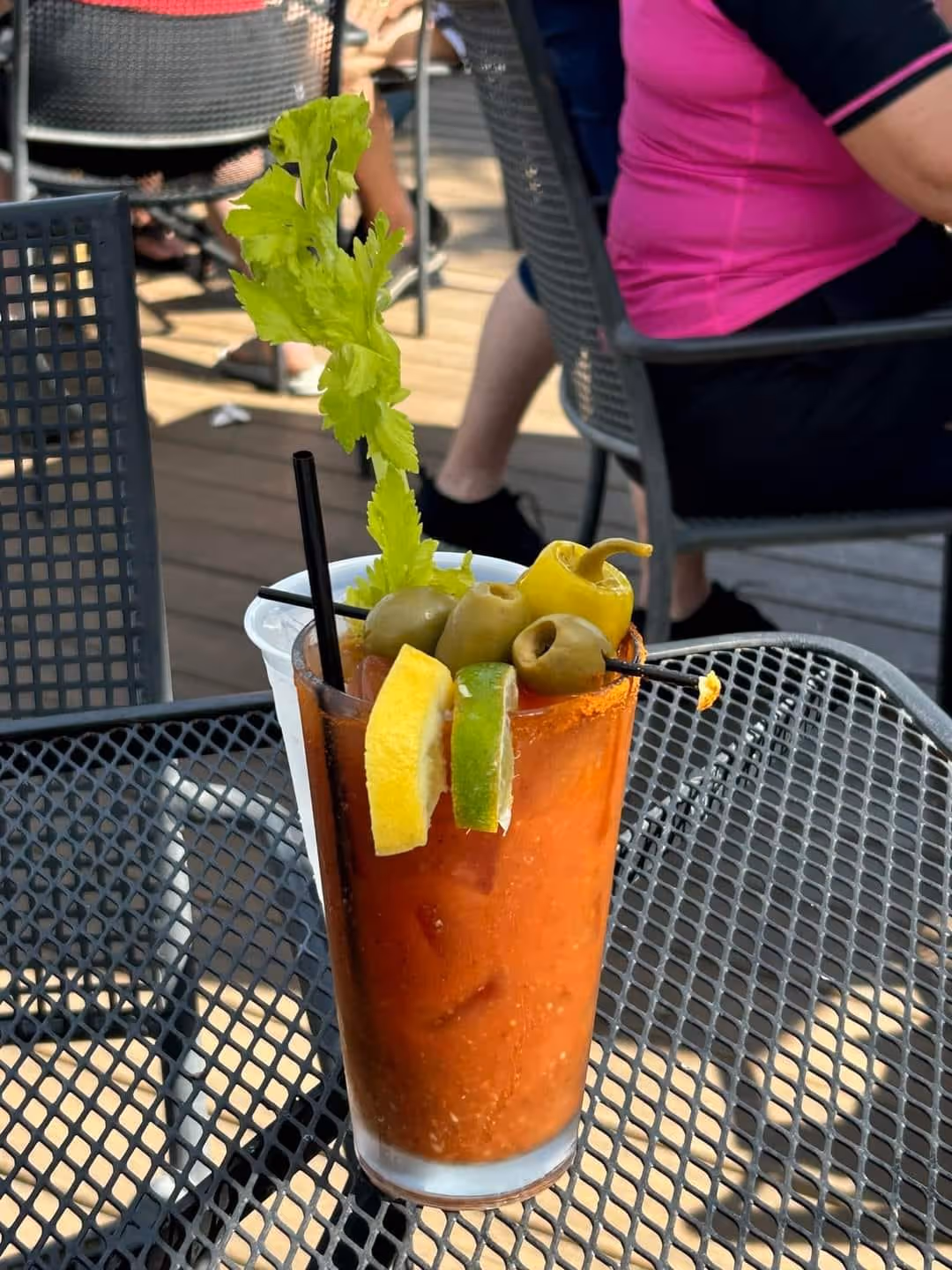 A Bloody Mary cocktail on a patio table, garnished with celery, olives, lemon, and lime slices. Nearby, people are seated at metal chairs, creating a relaxed and casual atmosphere.