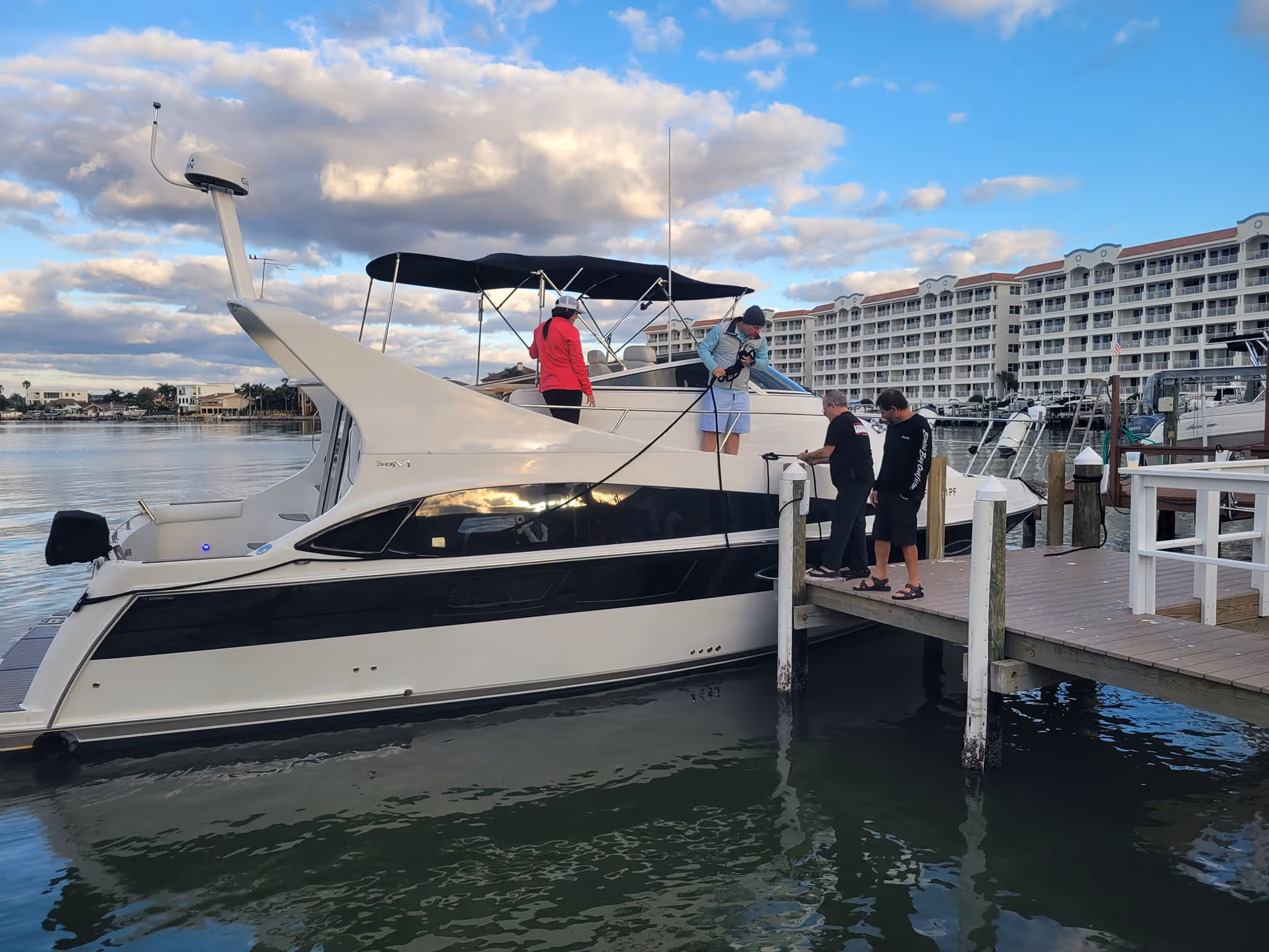 A group of people prepares a white motor yacht for departure from a wooden dock. The backdrop features a calm marina and a large hotel under a partly cloudy sky.