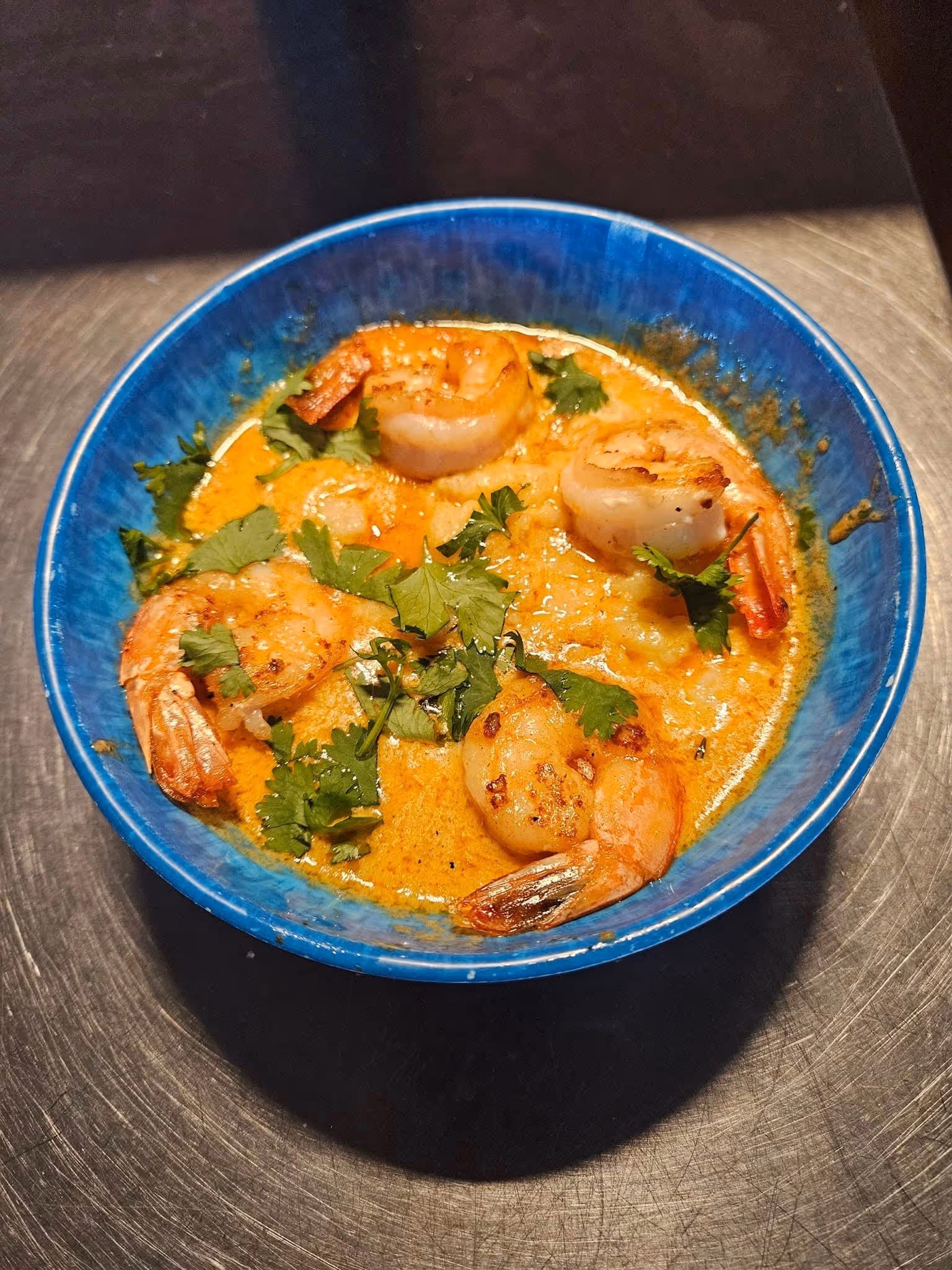 A blue bowl of creamy, golden shrimp curry garnished with fresh cilantro. The dish appears appetizing and warmly presented on a dark surface.