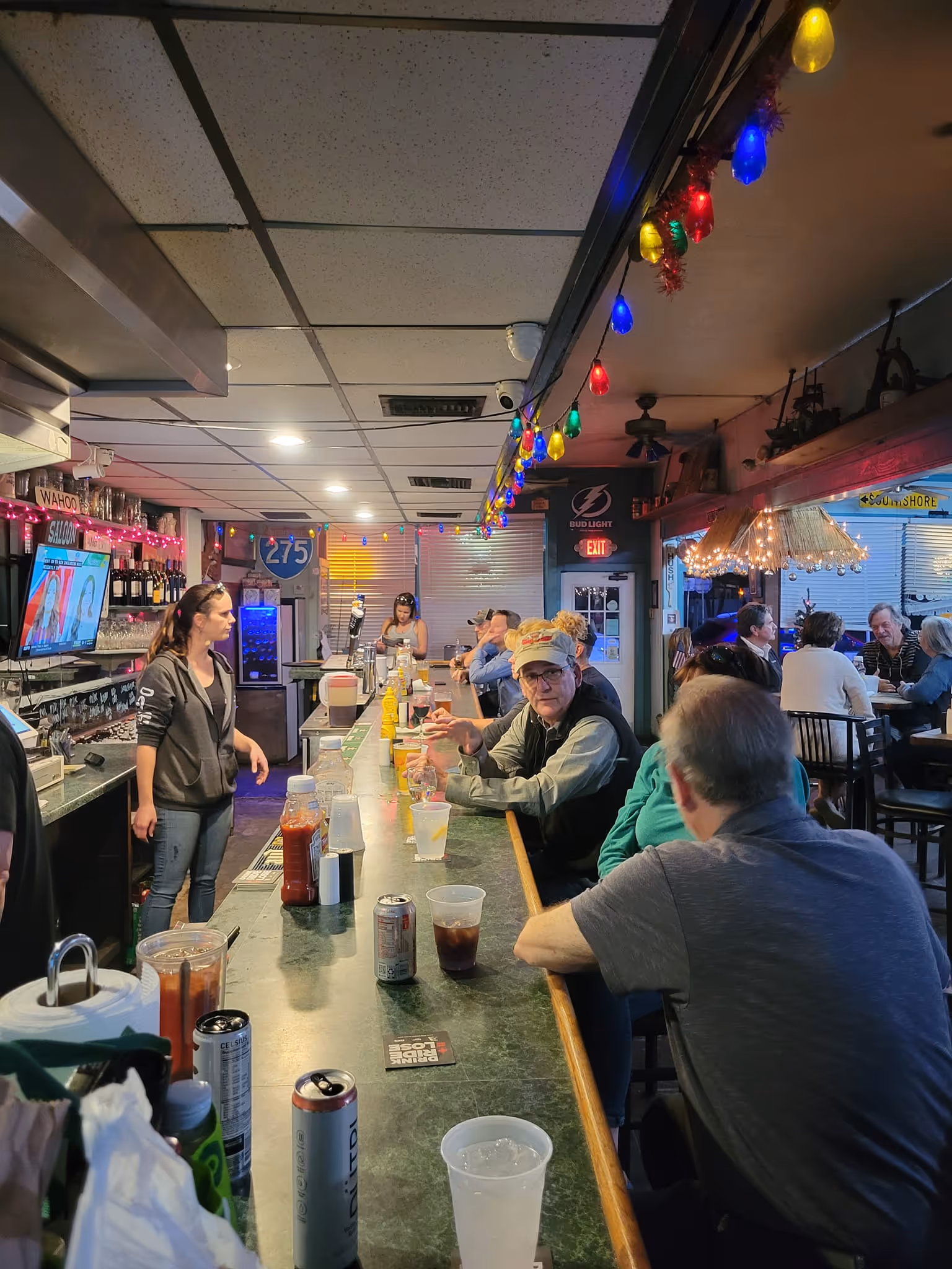A lively bar scene with people seated along a counter. A bartender serves drinks, with colorful lights hanging overhead, creating a festive and vibrant atmosphere.