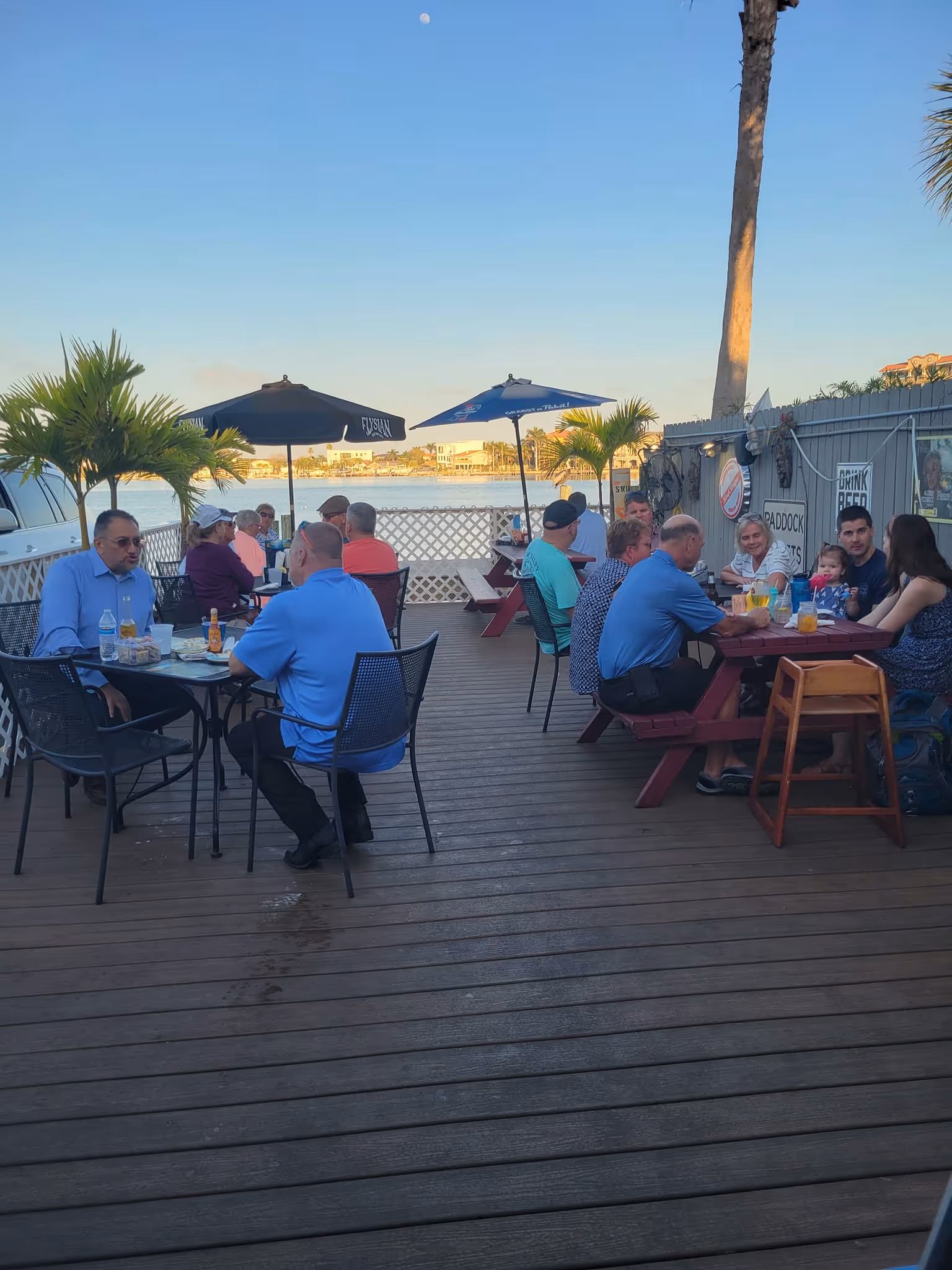 Outdoor restaurant scene on a wooden deck with people dining at tables under umbrellas. Blue sky and palm trees create a relaxed, summery atmosphere.