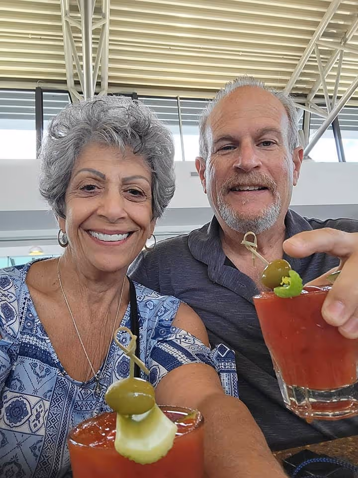 A smiling couple sits at a table with Bloody Mary cocktails garnished with olives and lime. The setting appears to be a bright, airy café.