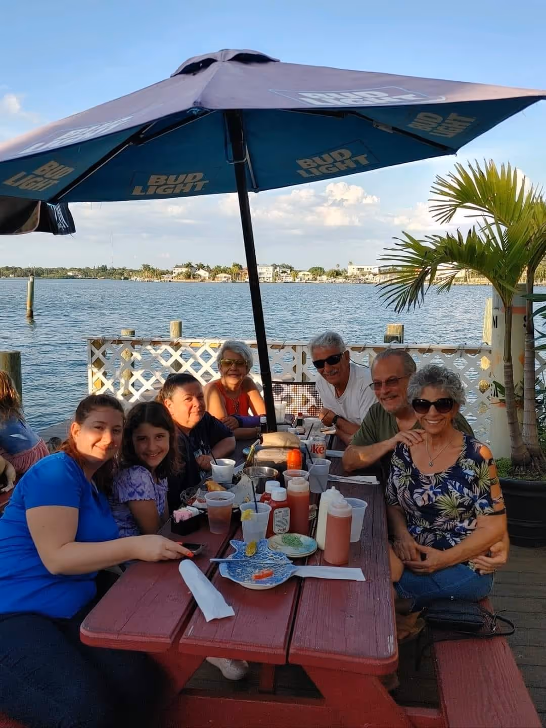 A group of people sit happily around a red picnic table on a sunny deck by the water. A large umbrella provides shade; palm trees and calm water are in the background.