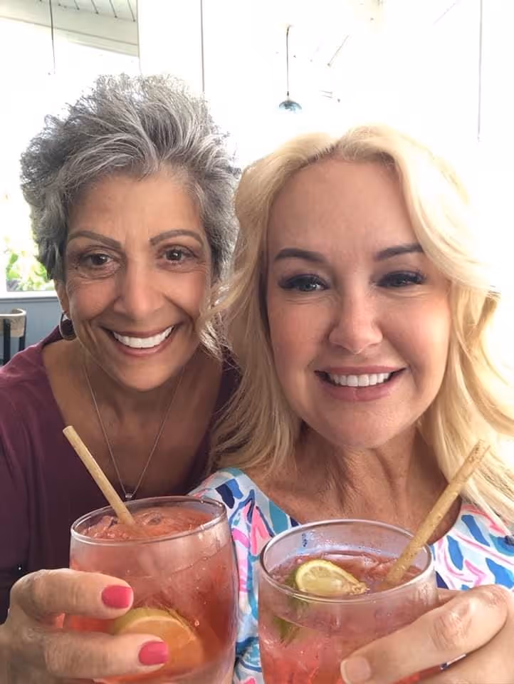 Two women smiling and holding pink drinks with lime slices in a bright room. They convey a joyful, relaxed mood, suggesting a moment of celebration.