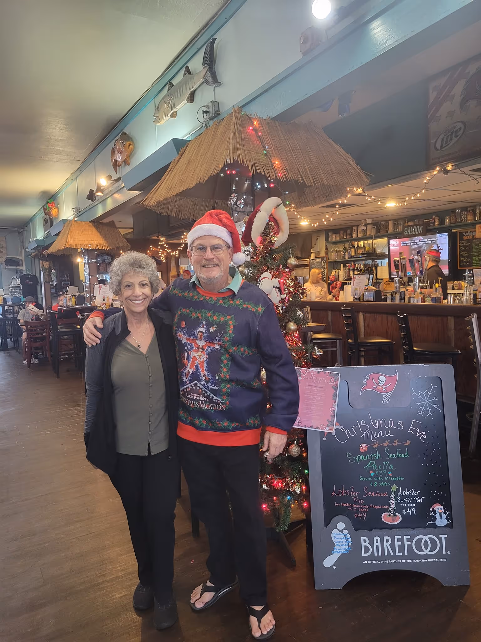 A smiling couple poses in a festive, tropical-themed restaurant. The man wears a Santa hat and Christmas sweater. A decorated Christmas tree and menu board are nearby.