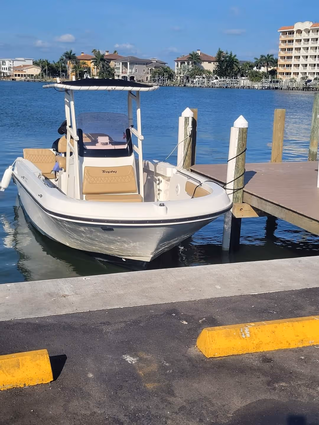 A small white boat with a canopy is docked by a wooden pier on calm blue water. Background shows waterfront homes and a clear sky. Peaceful setting.