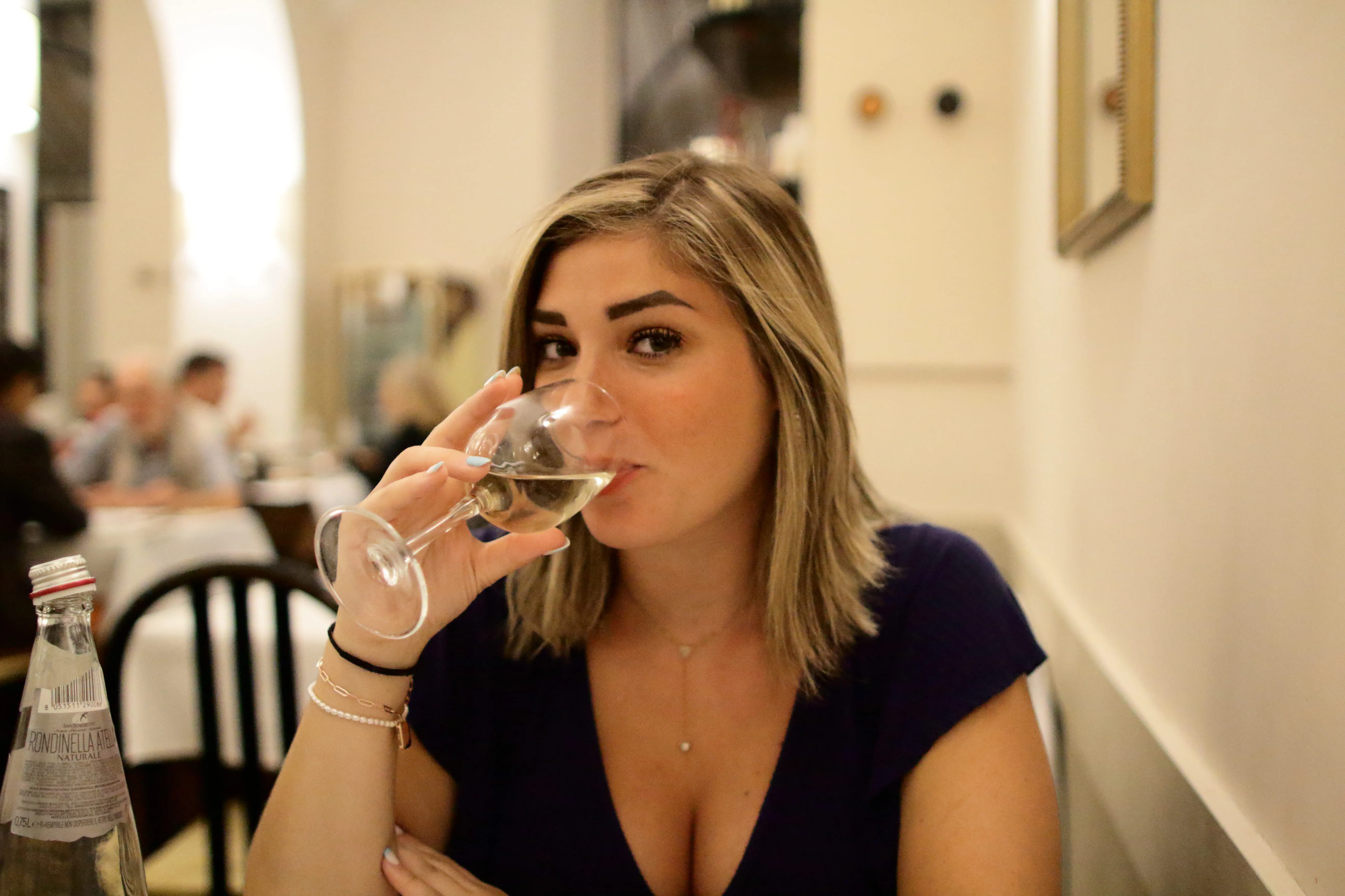Woman with blonde hair in dark shirt drinking white wine in a softly lit restaurant.