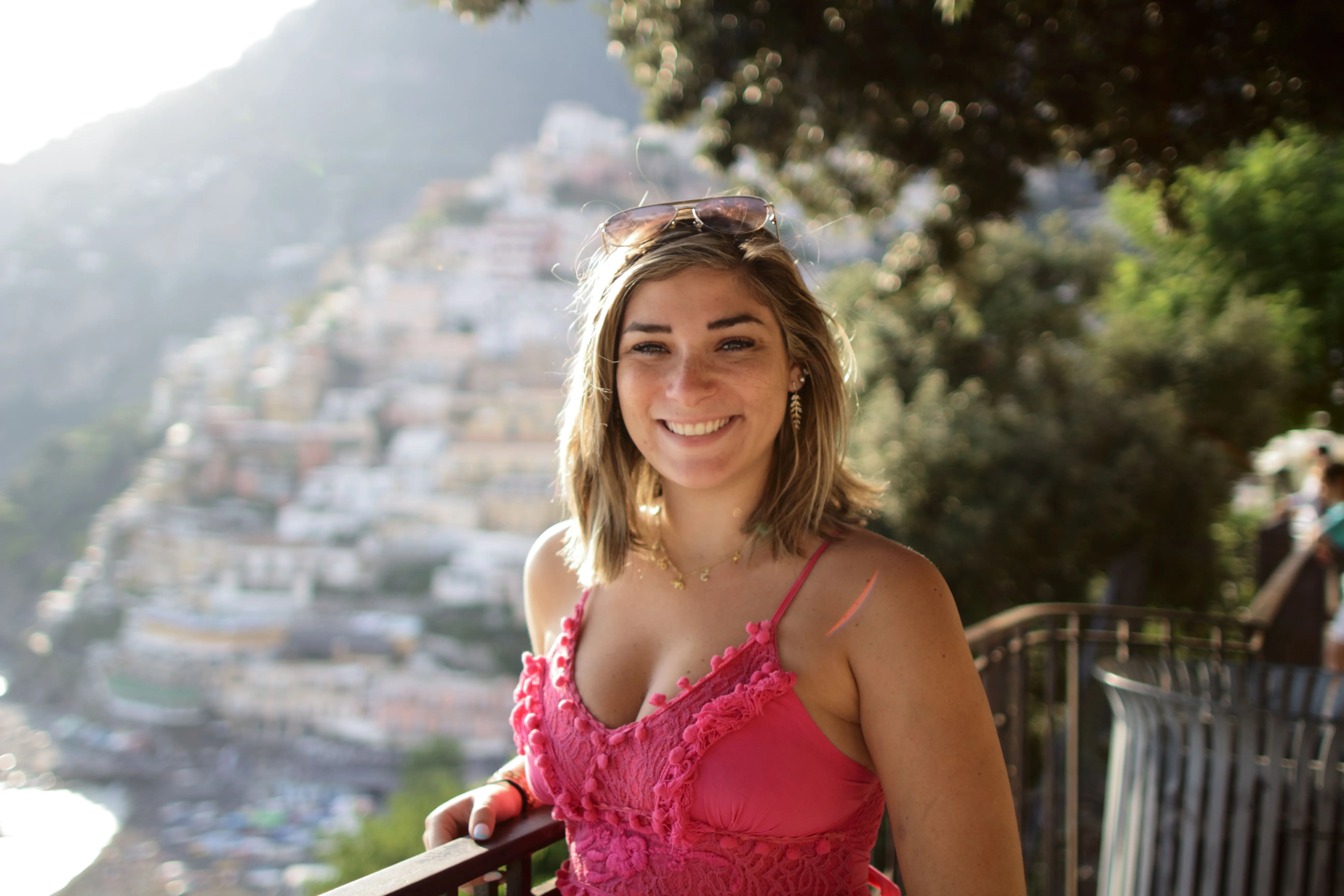 Smiling woman in pink dress with sunglasses on her head stands by railing with hillside town in background.
