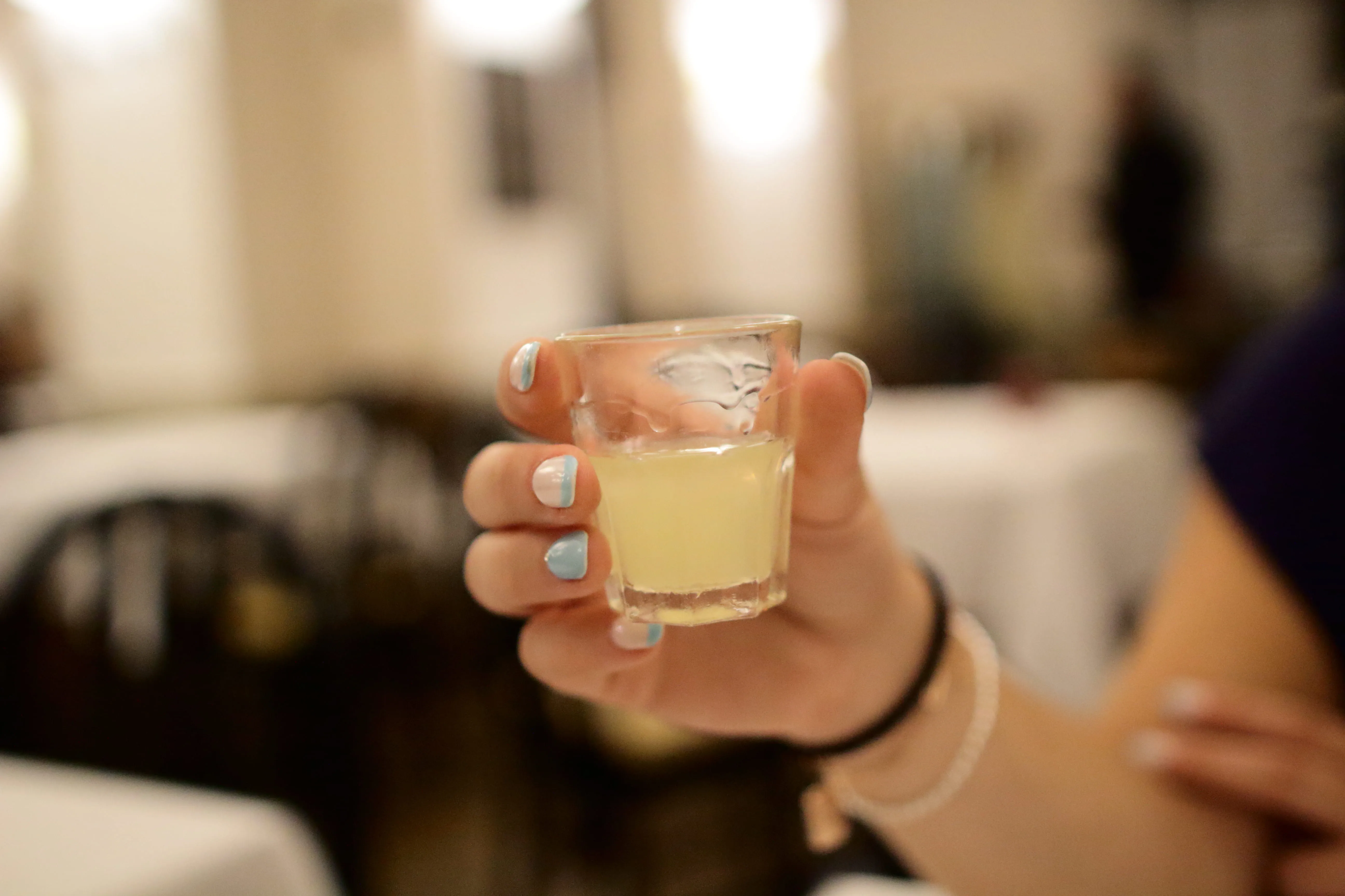 Hand with blue and white nail polish holding a small glass with pale yellow liquid.