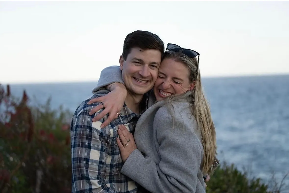 Smiling couple hugging near a body of water with greenery in the background.